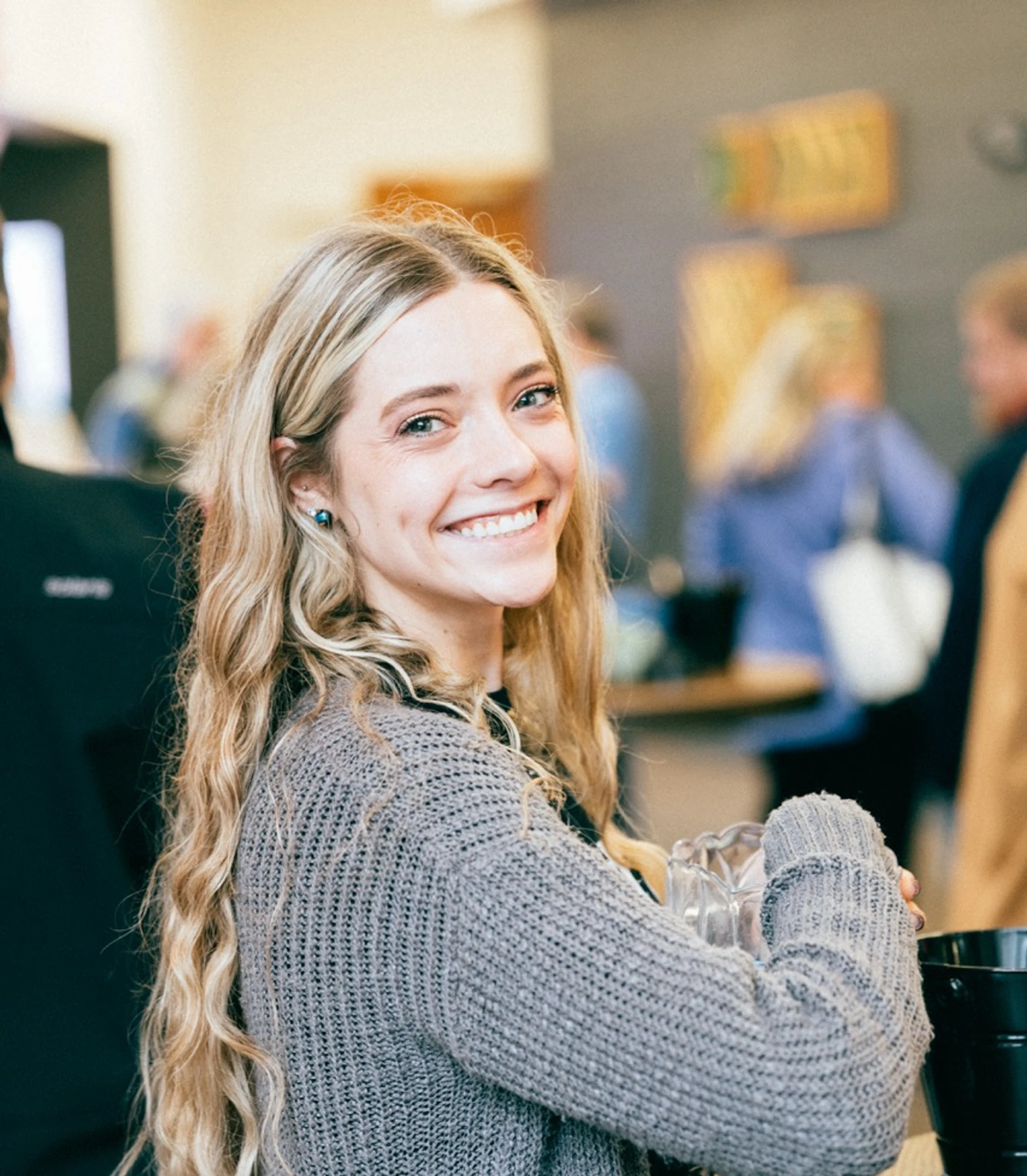 A young woman with long, wavy blonde hair and a friendly smile looks at the camera. She wears a gray knit sweater and holds a glass pitcher indoors.