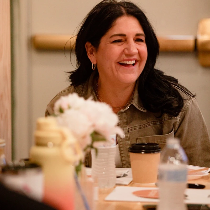 A candid shot of a woman with dark hair laughing while sitting at a table with flowers, a coffee cup, and water bottles during a workshop or meeting.