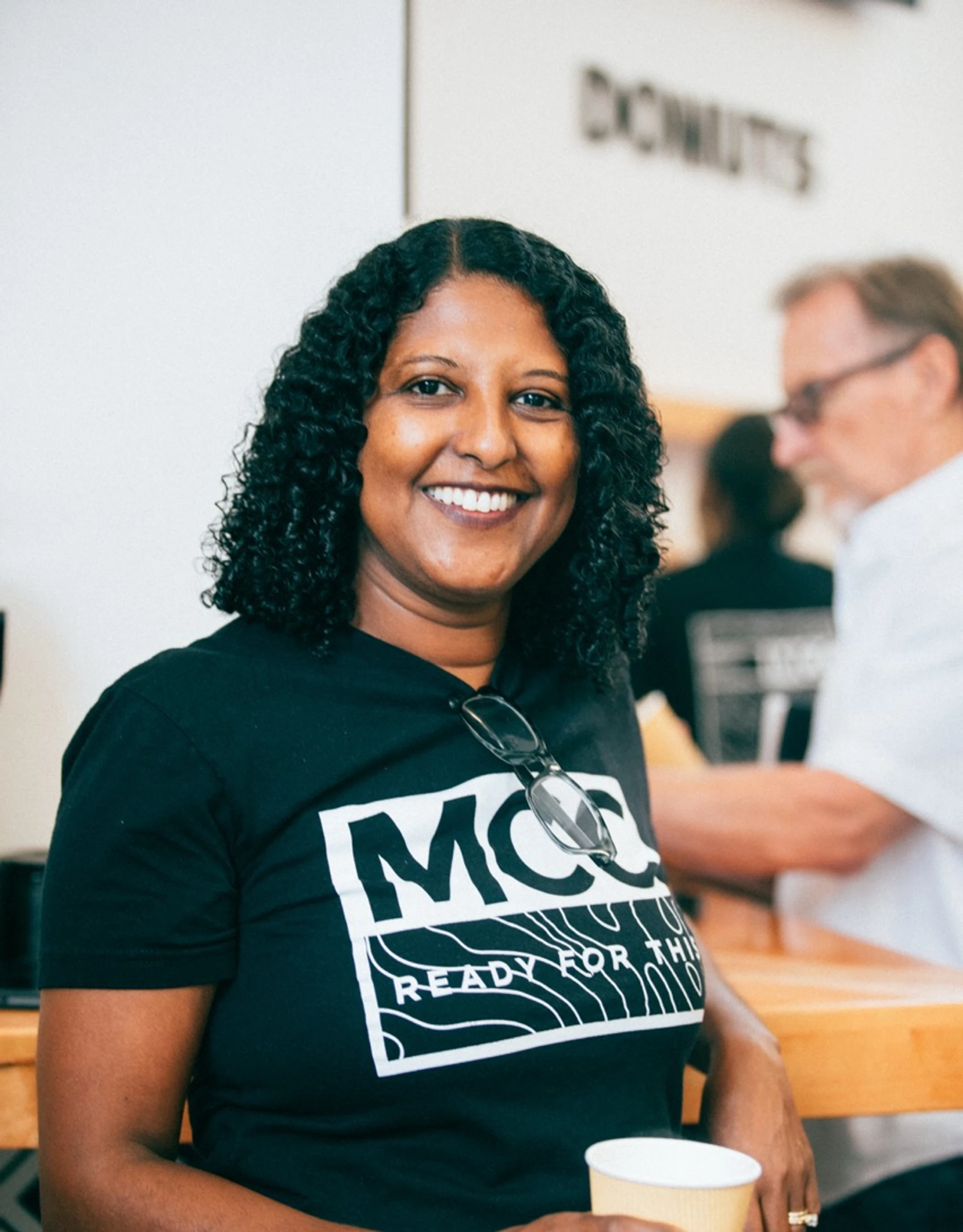 A woman with dark curly hair smiles warmly at the camera. She's wearing a black t-shirt that says 'MCC Ready For This' and holding a paper cup.