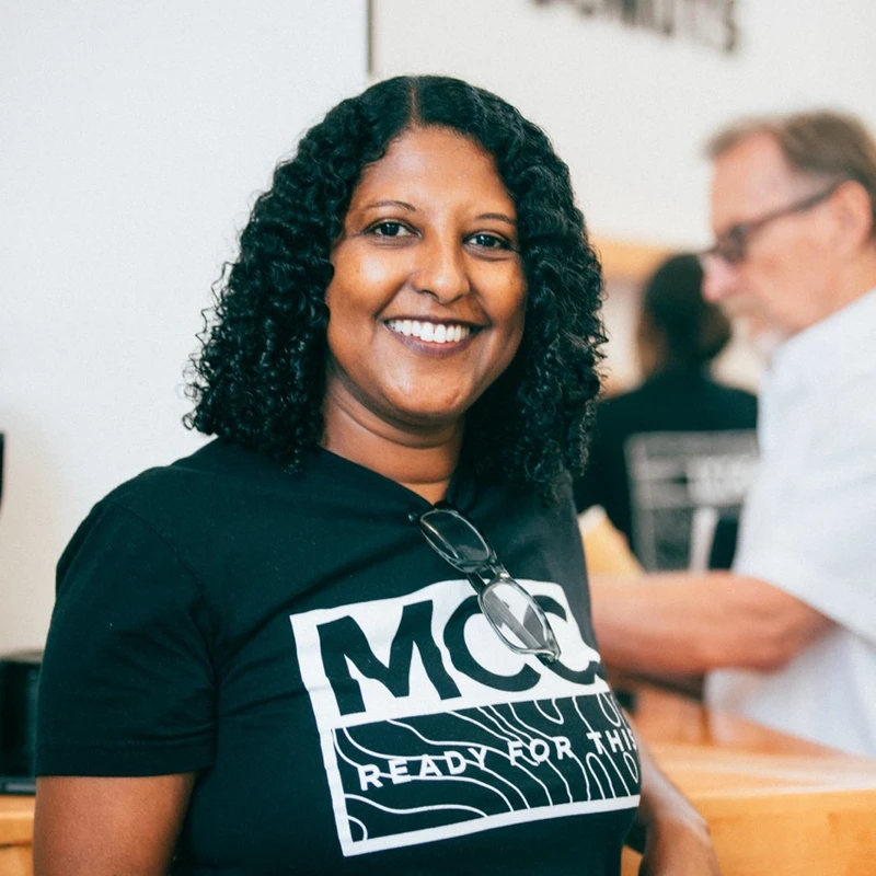 A woman with dark curly hair smiles warmly at the camera. She wears a black t-shirt with the text 'MCC READY FOR THIS' and holds a paper cup.