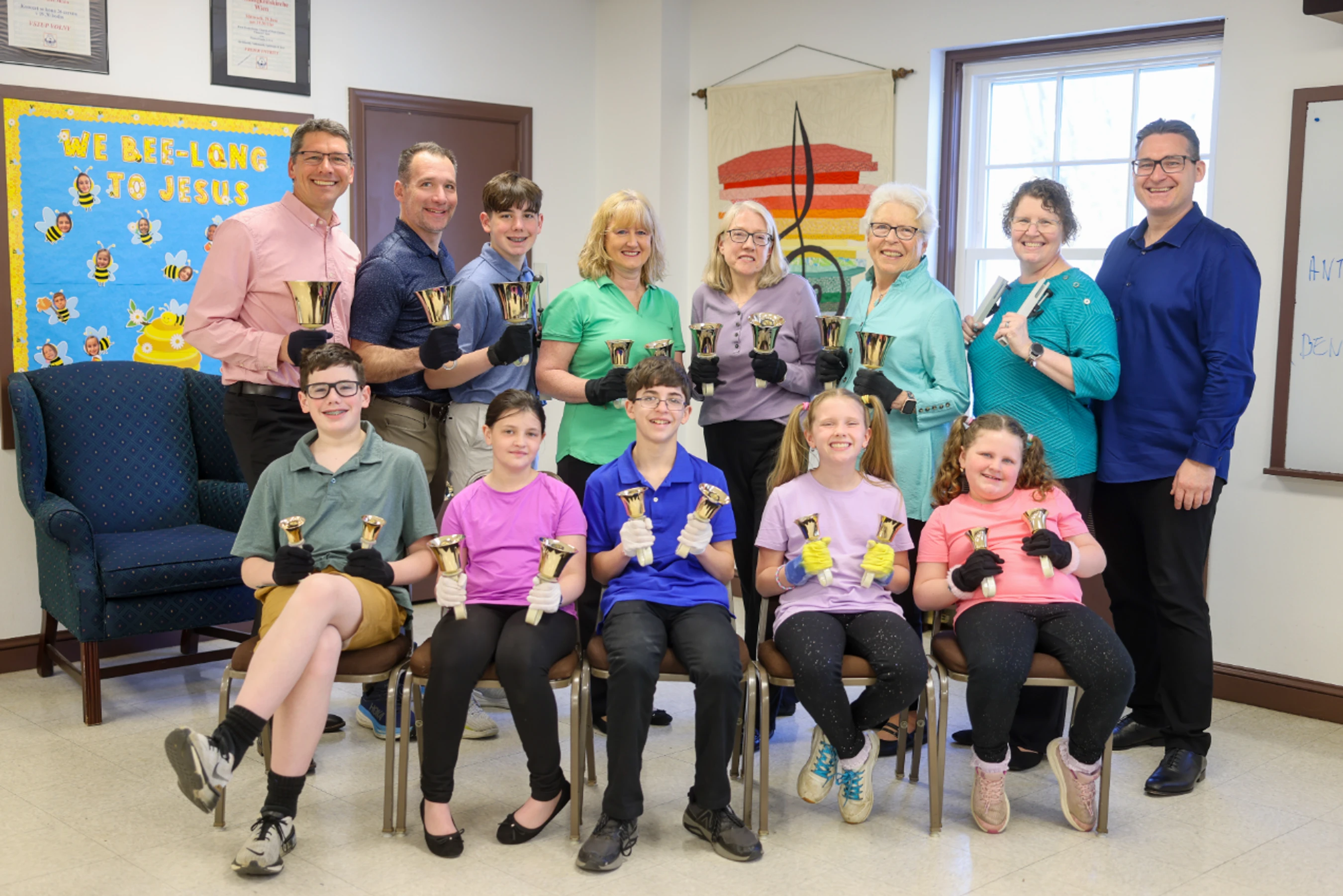 A group of smiling adults and children pose for a photo in a classroom. They are all wearing gloves and holding one or two golden handbells.