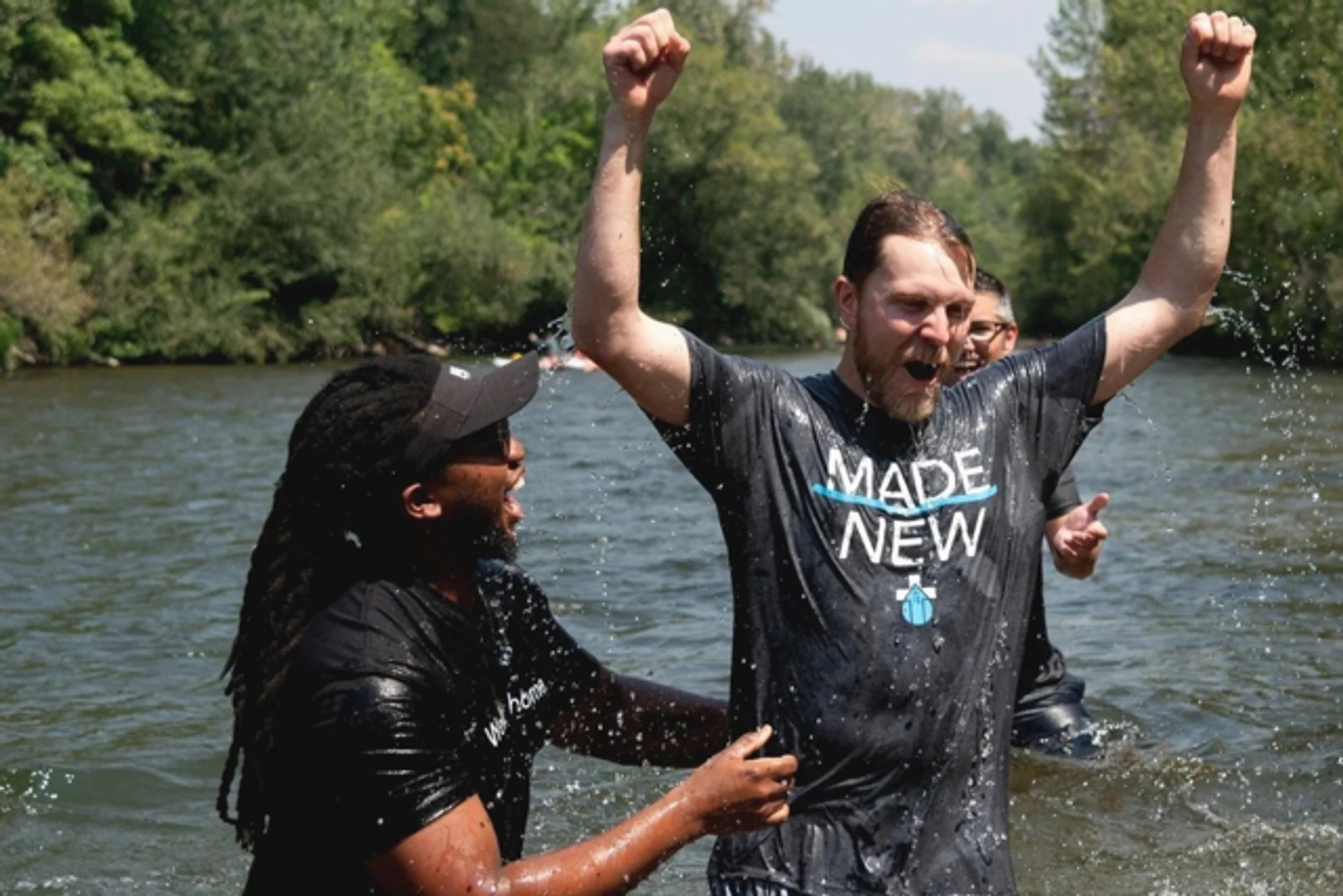 A man in a 'Made New' t-shirt raises his arms in celebration as another man baptizes him in a river, both smiling joyfully amidst splashing water.