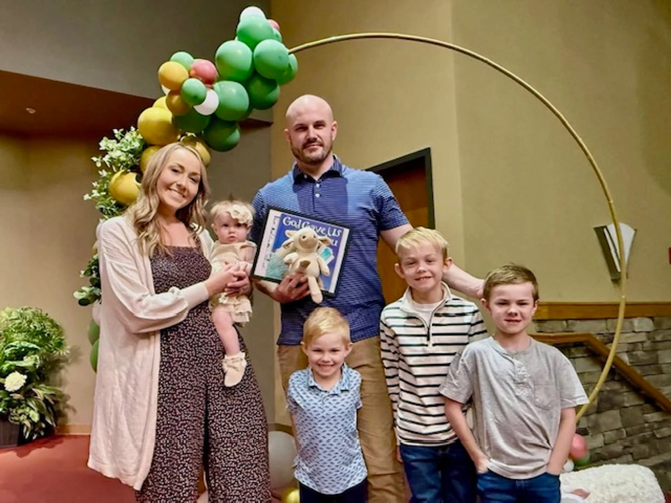 A man and woman pose with their four children, three boys and a baby girl, smiling in front of a green and yellow balloon arch during a celebration.
