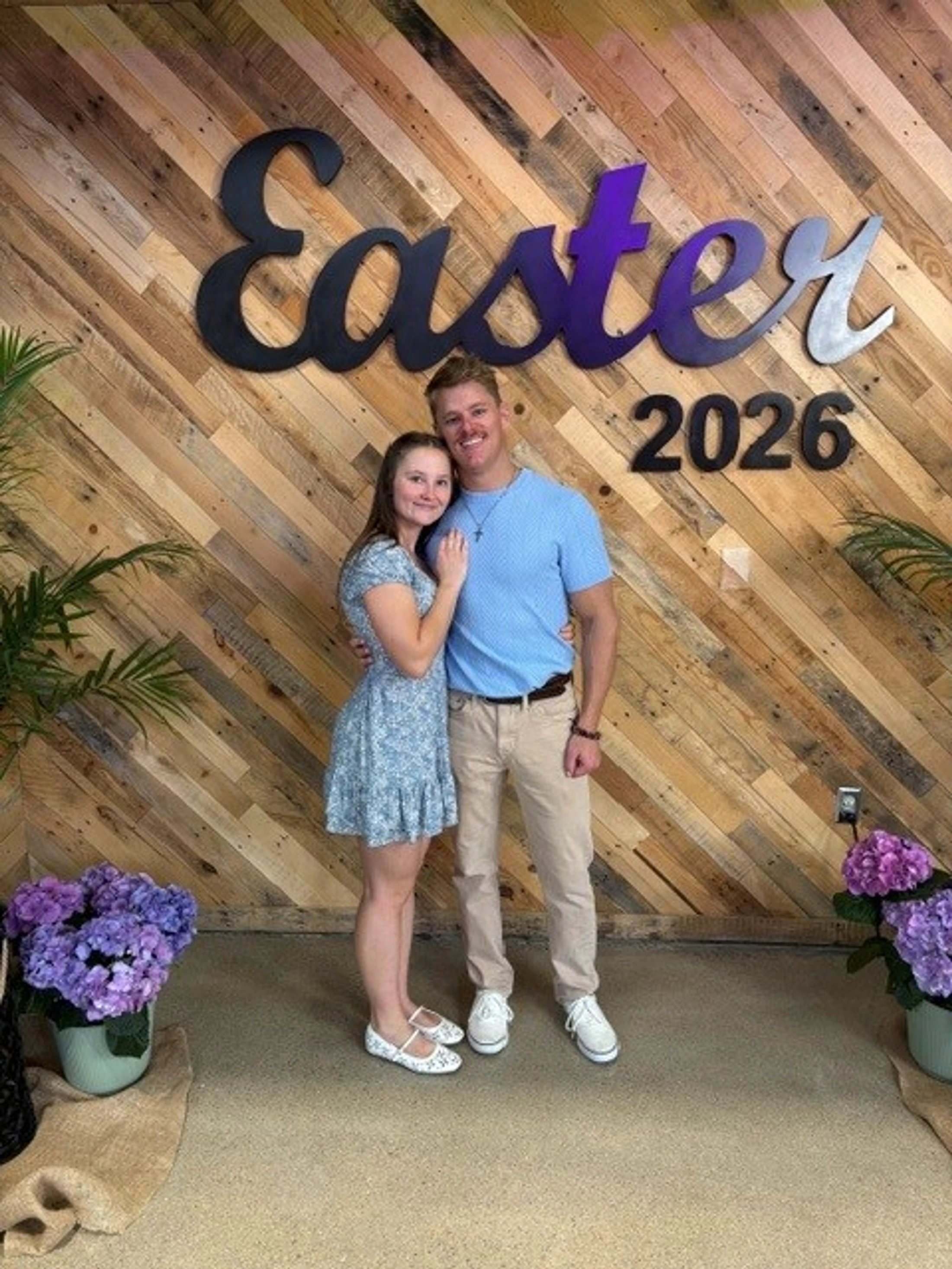 A smiling young couple stands in front of a rustic wooden wall with a large sign that says 'Easter 2026'. They are flanked by pots of purple flowers.