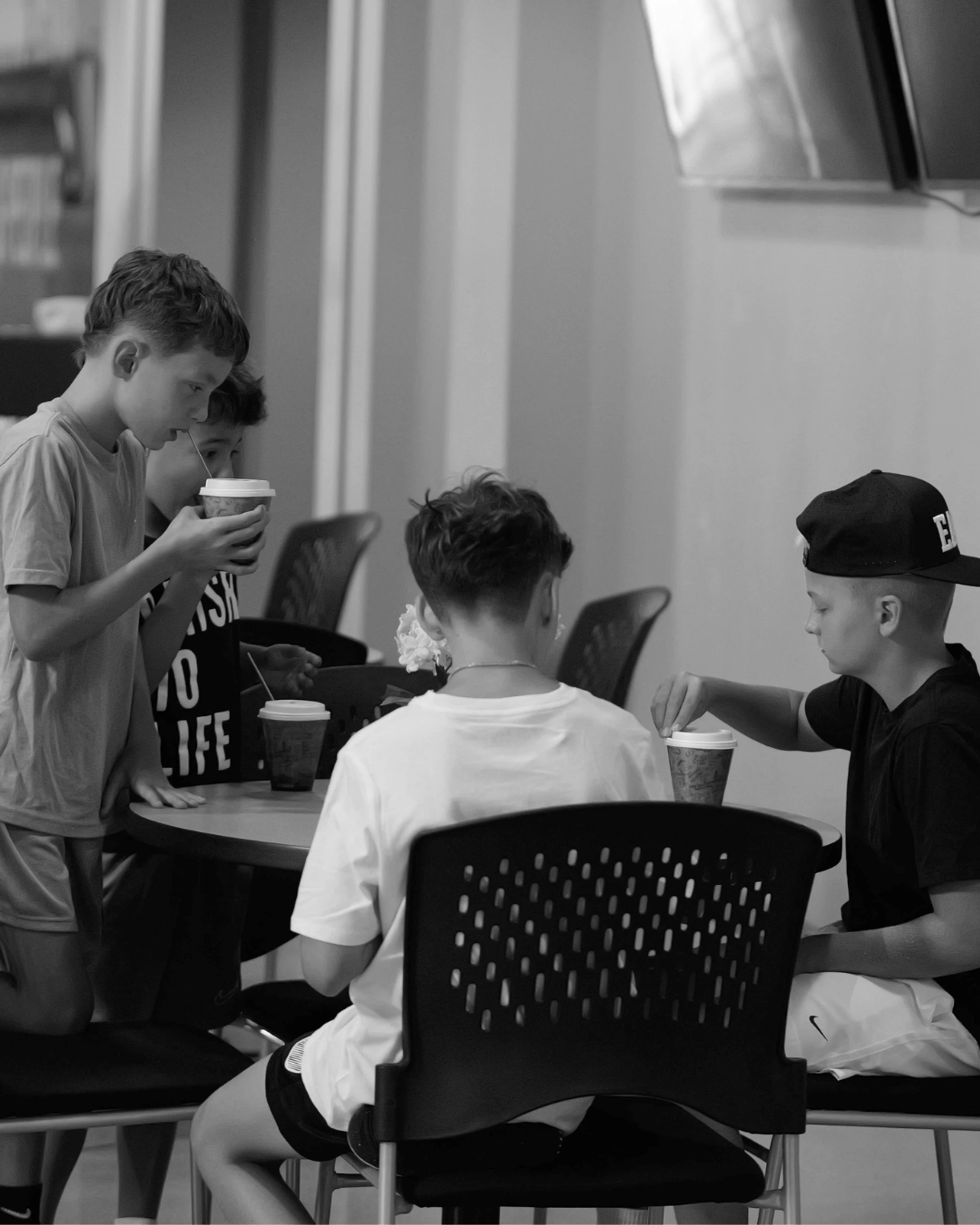 A black and white photo of four young boys gathered around a table, drinking from disposable cups with straws in a casual indoor setting.