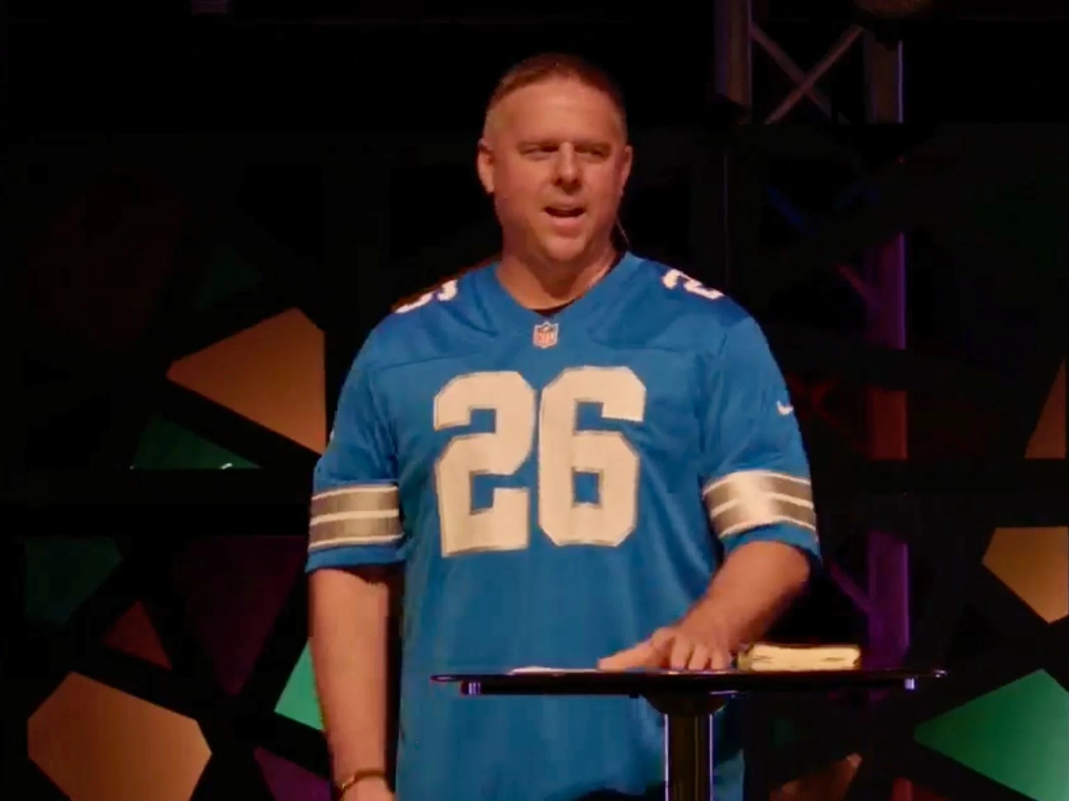 A man wearing a blue Detroit Lions football jersey with the number 26 stands behind a lectern on a stage, speaking.