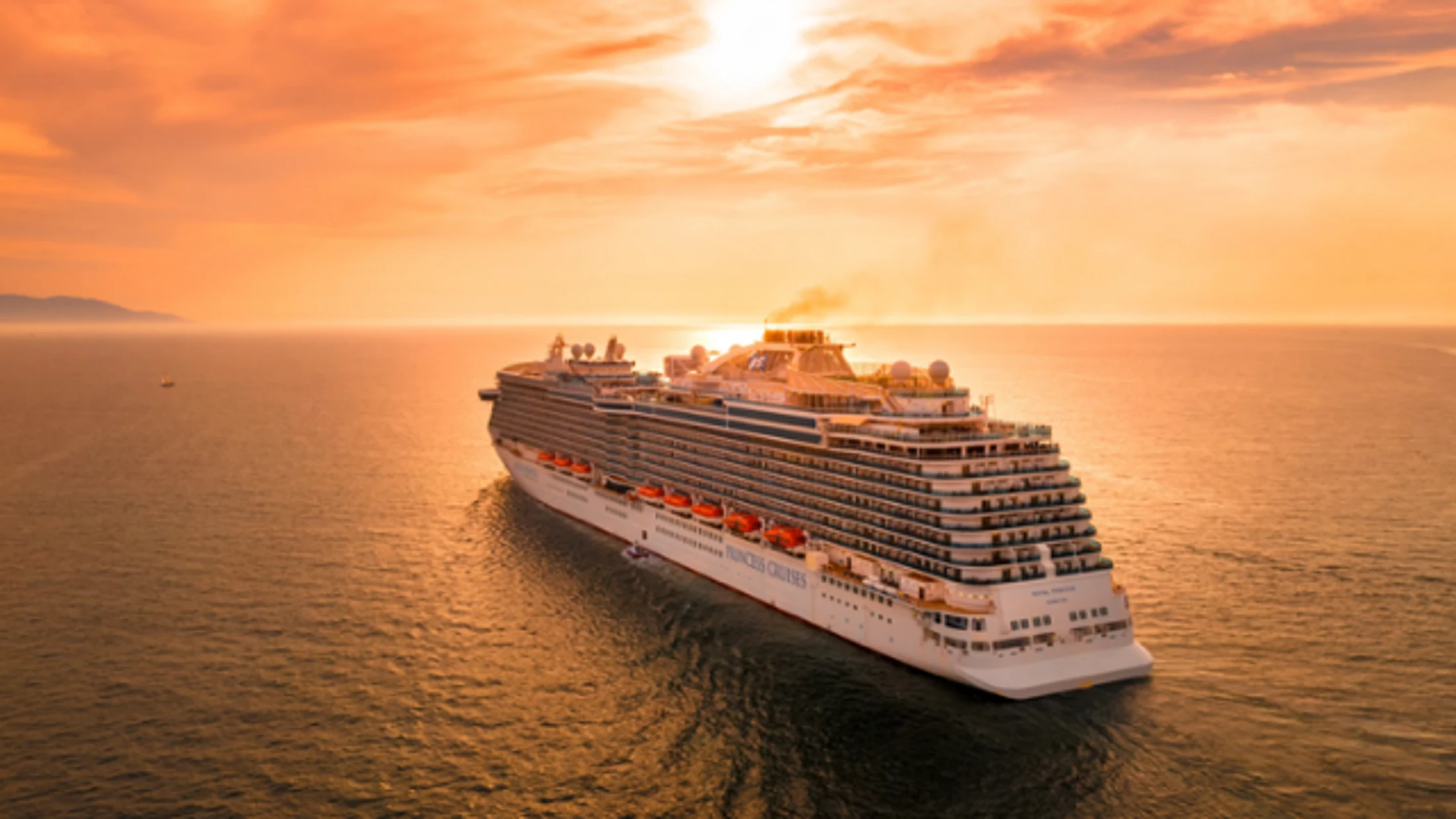 An aerial view of a large white Princess Cruises ship, the Royal Princess, sailing on a calm ocean during a brilliant orange and gold sunset.