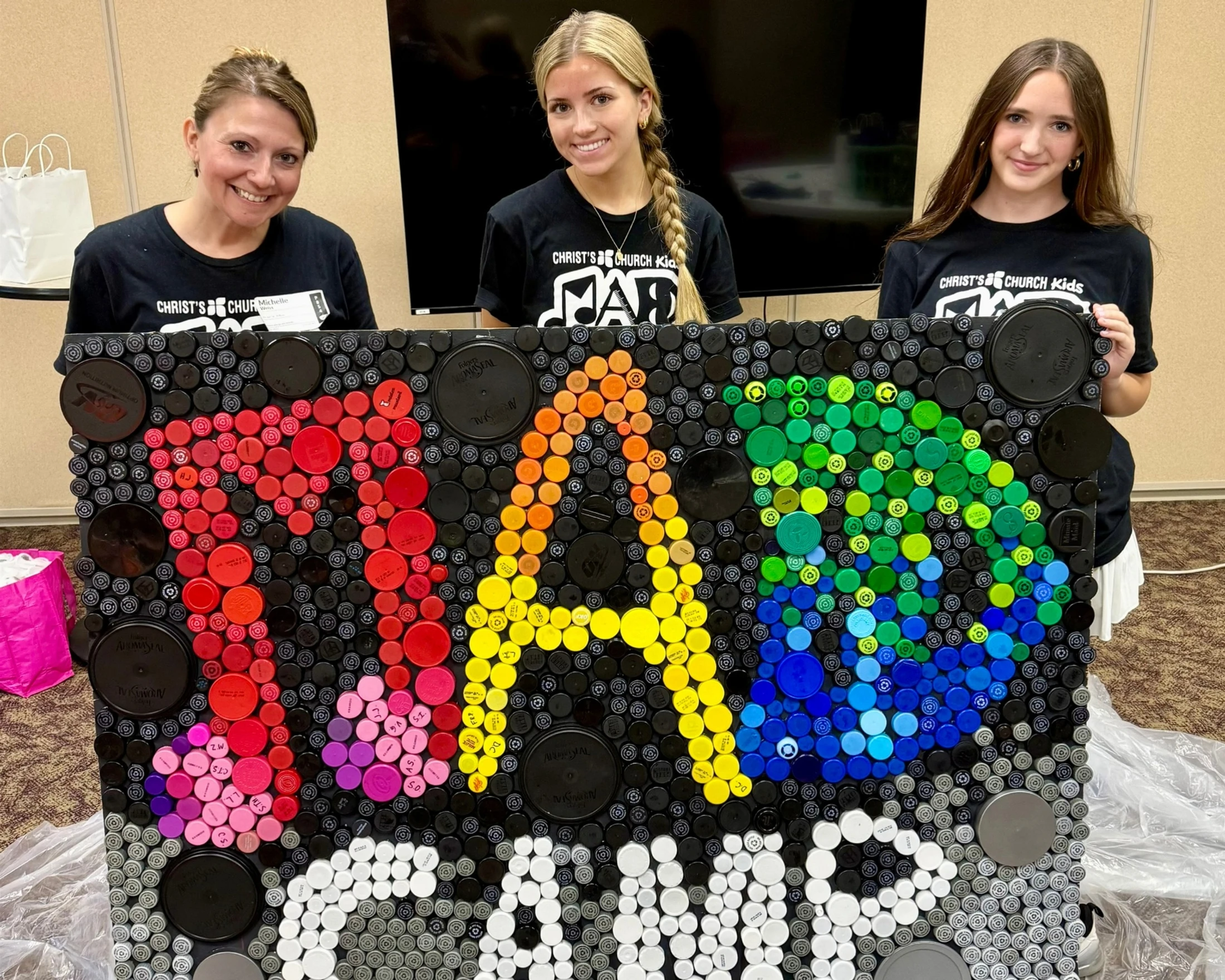 Three smiling women in matching black t-shirts hold a large art mosaic made from colorful recycled plastic bottle caps that spells out 'MAD CAMP'.