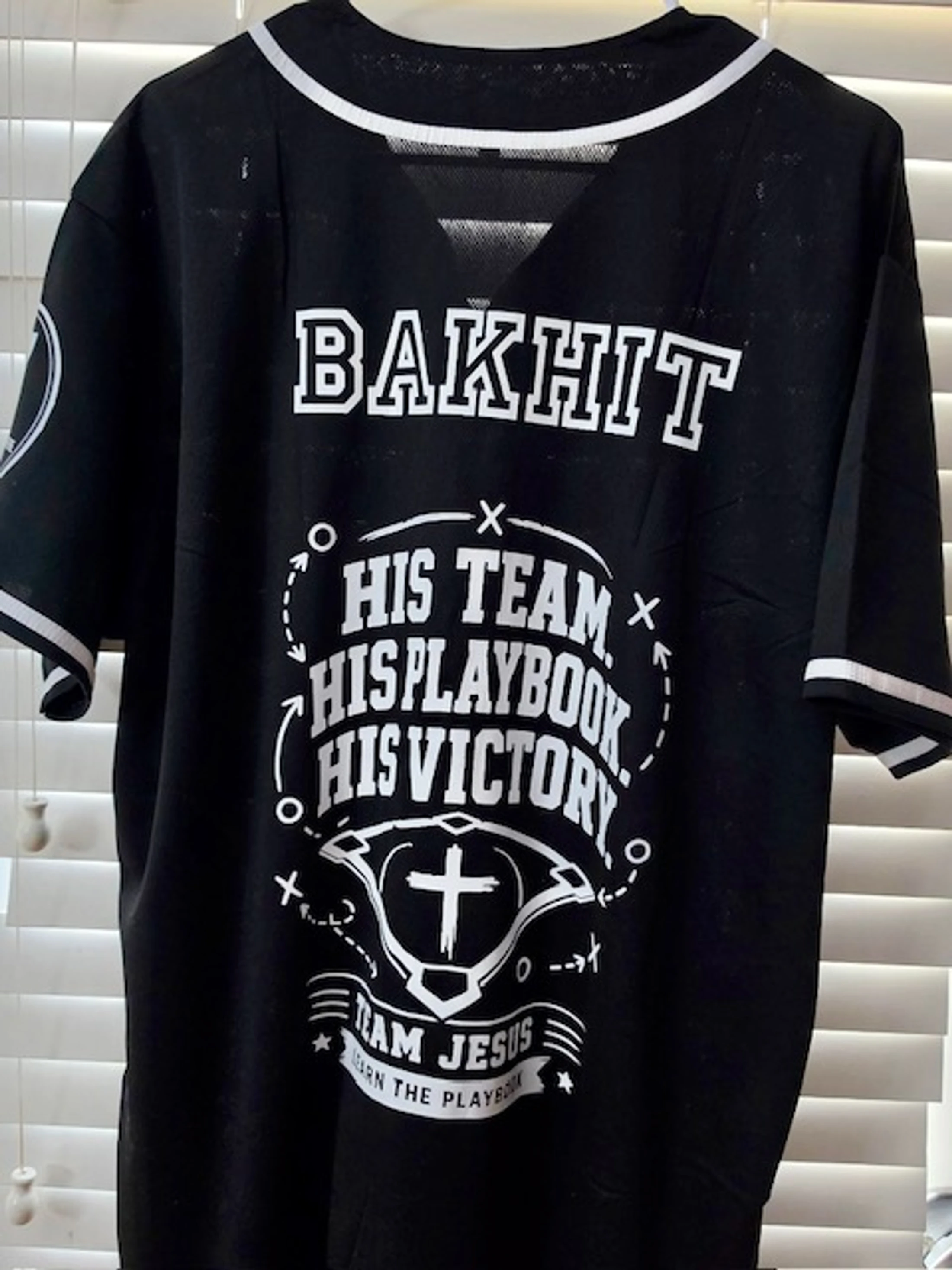The back of a black baseball-style jersey with 'BAKHIT' on the shoulders and a 'Team Jesus' graphic with a cross and football playbook diagrams.