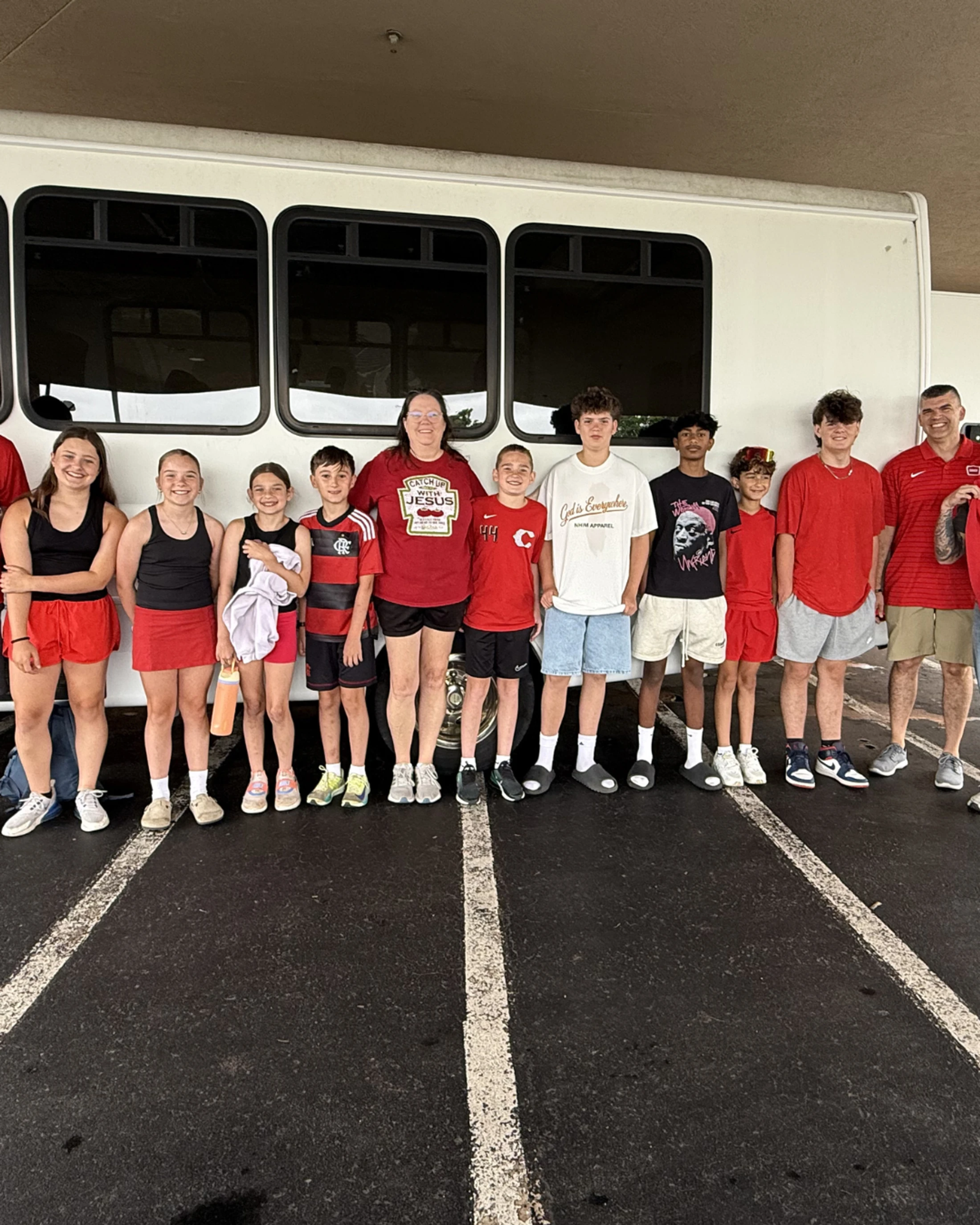 A group of children, teenagers, and two adults wearing mostly red shirts stand in a line and smile for a photo in front of a white bus in a parking lot.