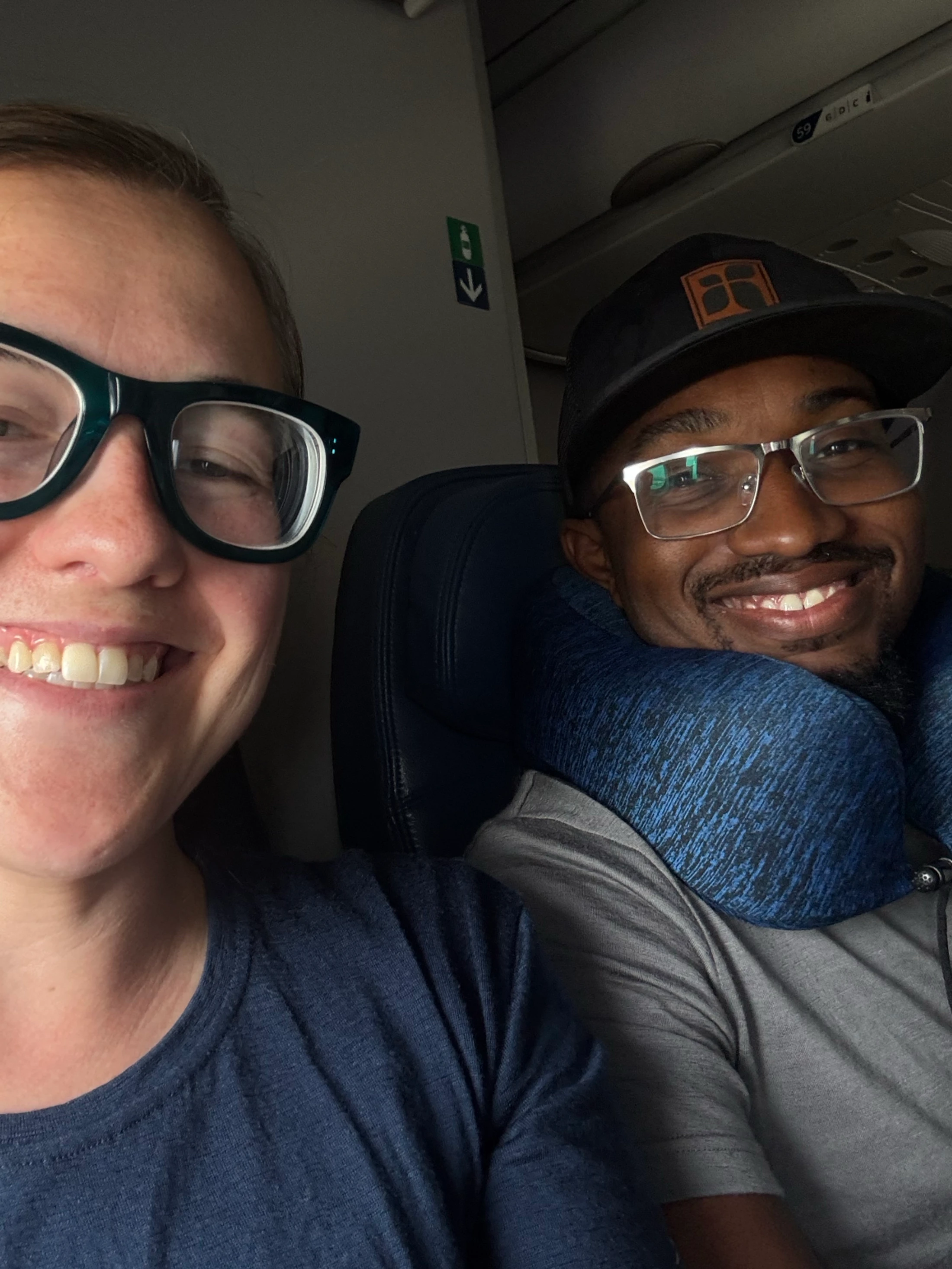 A close-up selfie of a smiling woman in glasses and a man in a hat and travel pillow, sitting side-by-side in their seats on an airplane.