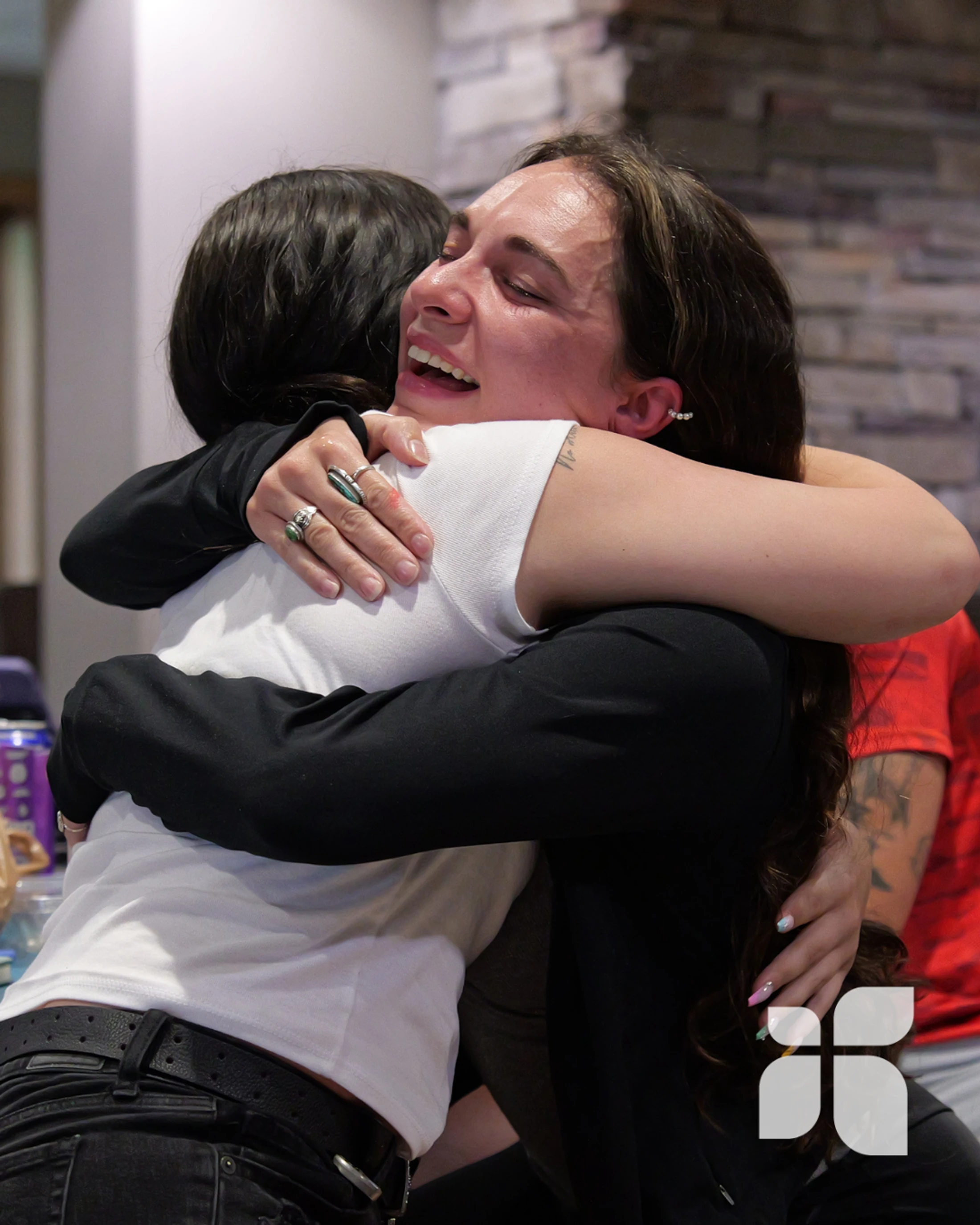 A woman with long brown hair smiles with her eyes closed while warmly hugging another person. She wears a black jacket over a white shirt and has several rings.