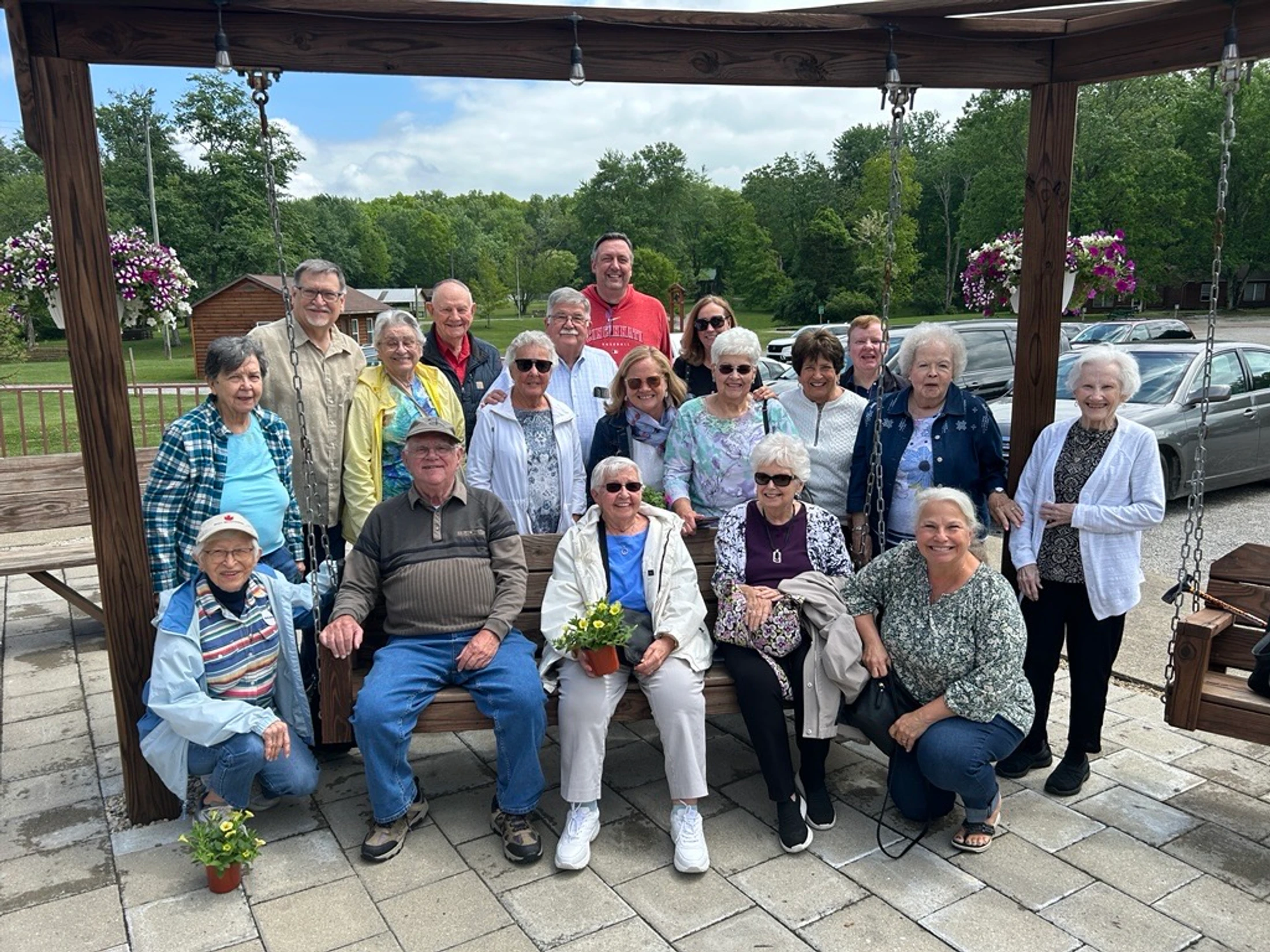 A large group of smiling seniors gathers for a photo on an outdoor patio. They are sitting and standing under a wooden pergola, with trees in the background.
