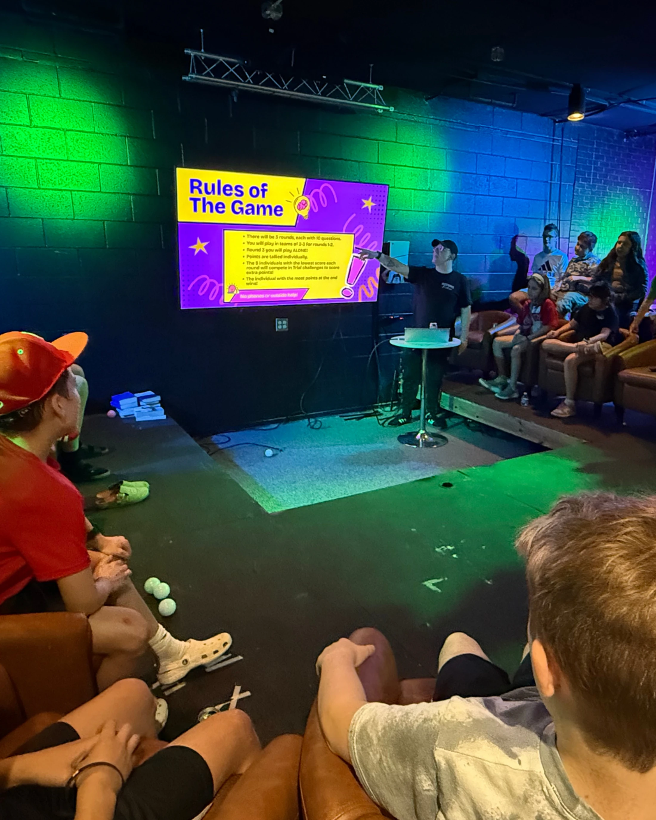 A man on a small stage points to a screen that says "Rules of The Game" while an audience of young people sits and watches in a room with green and blue lights.