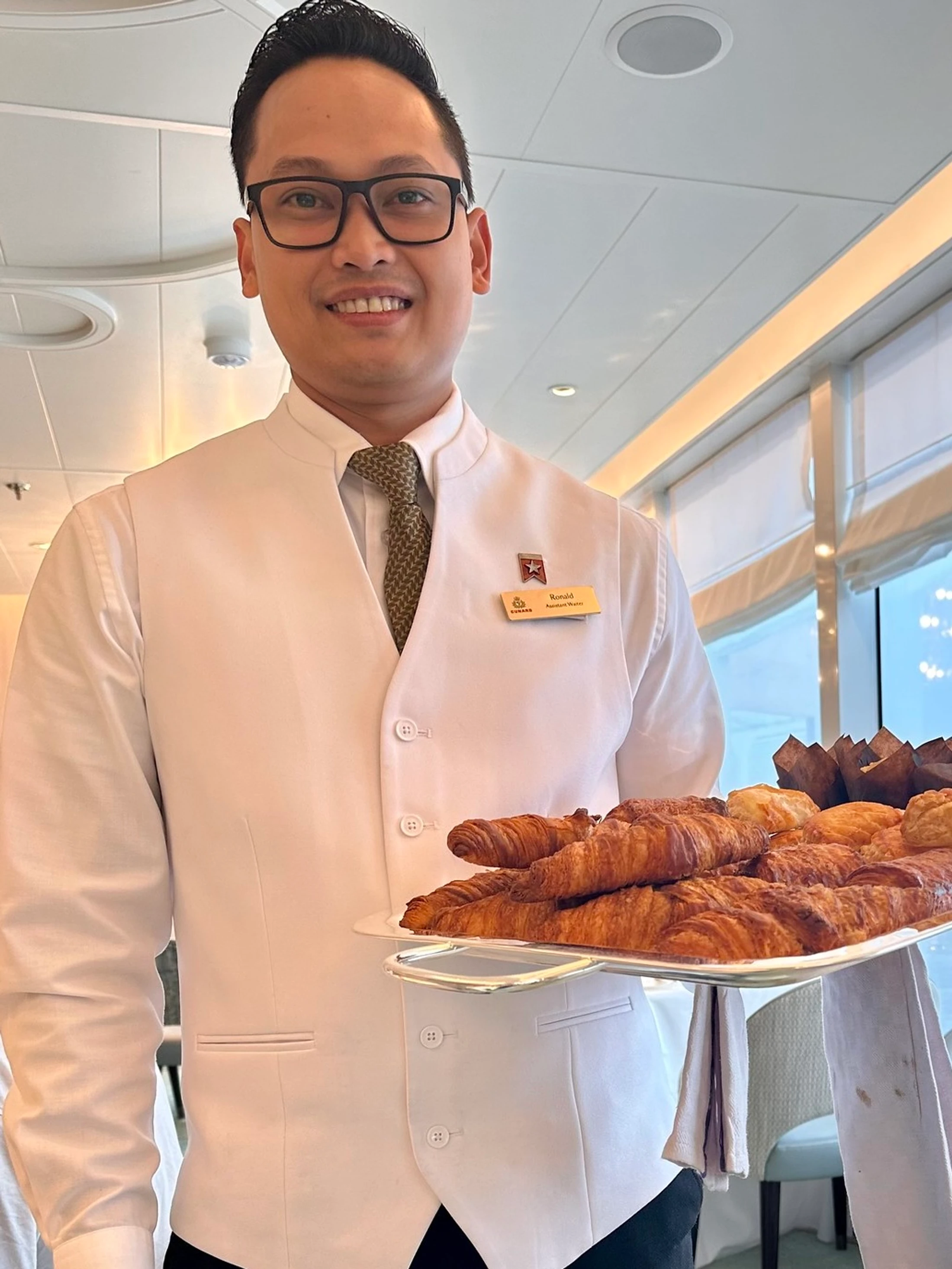 A smiling waiter in a white uniform and glasses holds a silver tray piled high with fresh croissants, muffins, and other breakfast pastries.