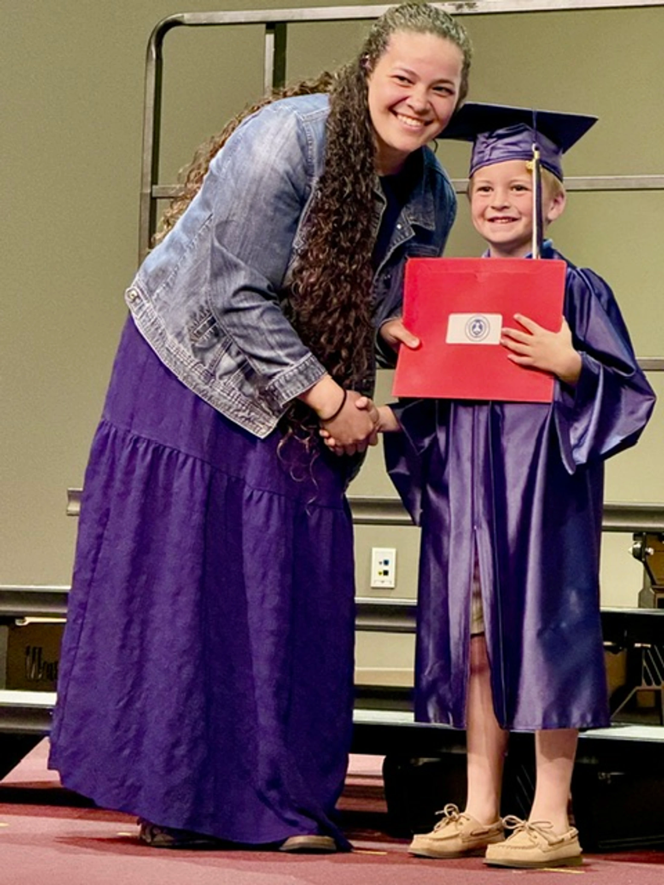 A woman with long curly hair shakes hands with a young boy wearing a purple cap and gown and holding a red diploma folder at his graduation ceremony.