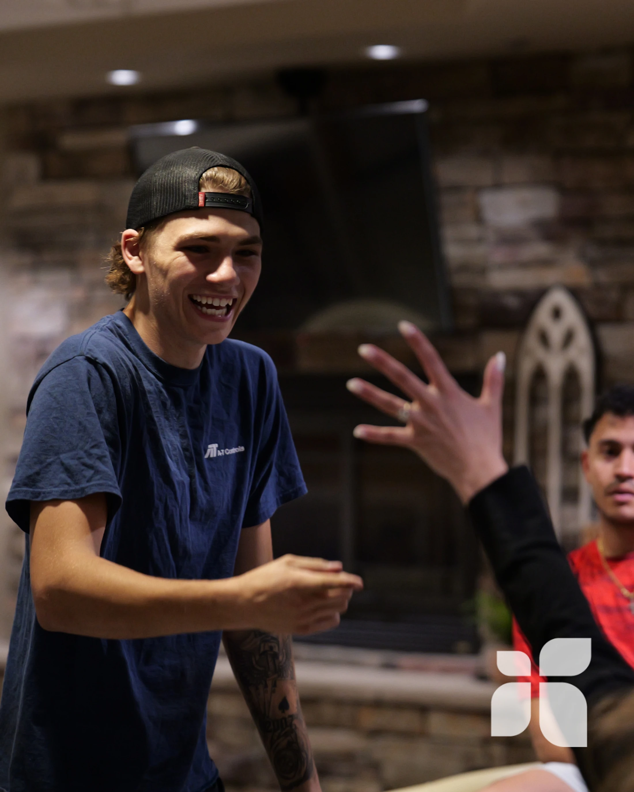 A young man in a blue t-shirt and backward baseball cap laughs while interacting with a friend whose hand is visible at an indoor social gathering.