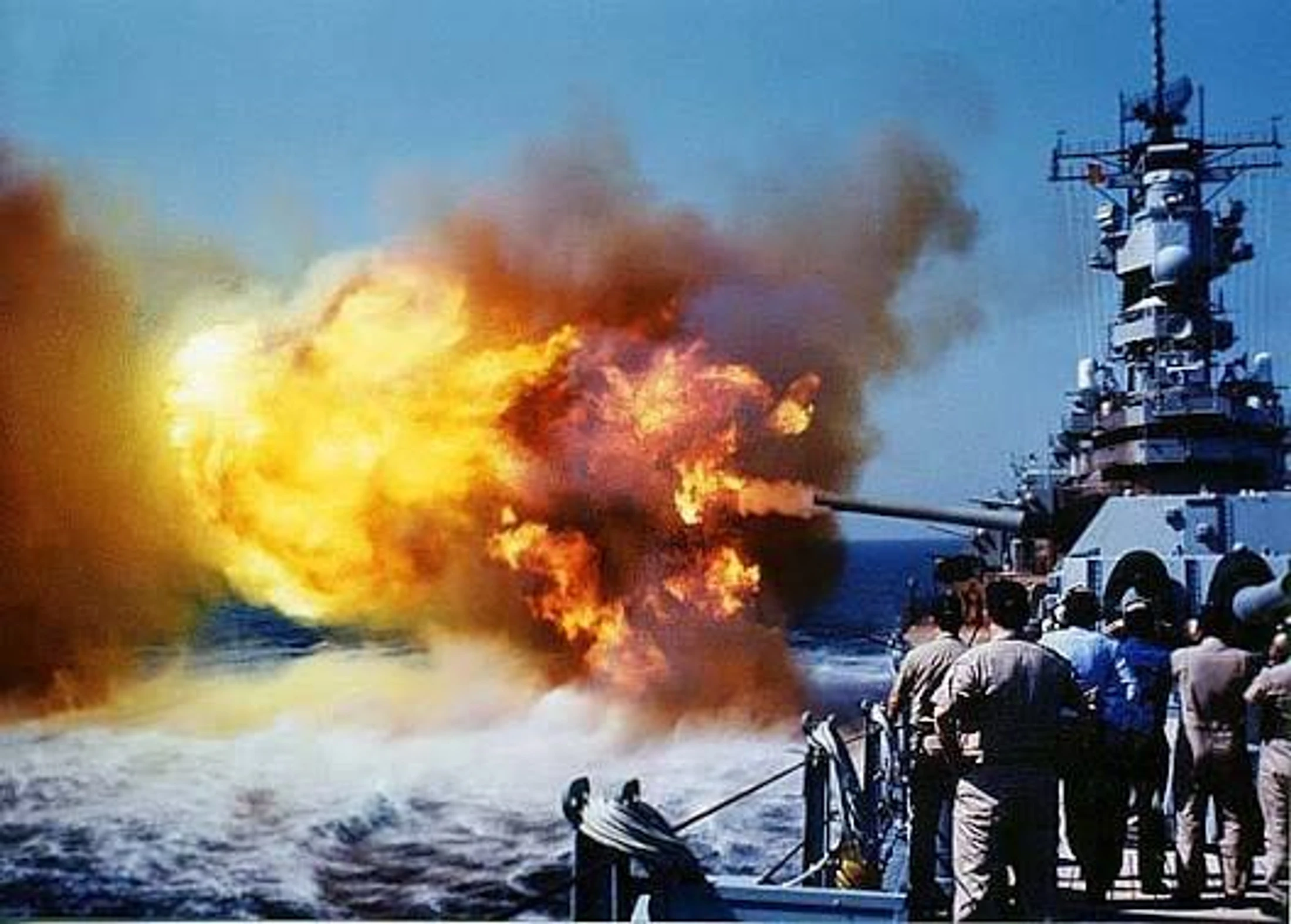A large naval warship fires a cannon, creating a massive fireball and smoke cloud over the ocean as sailors watch from the deck.