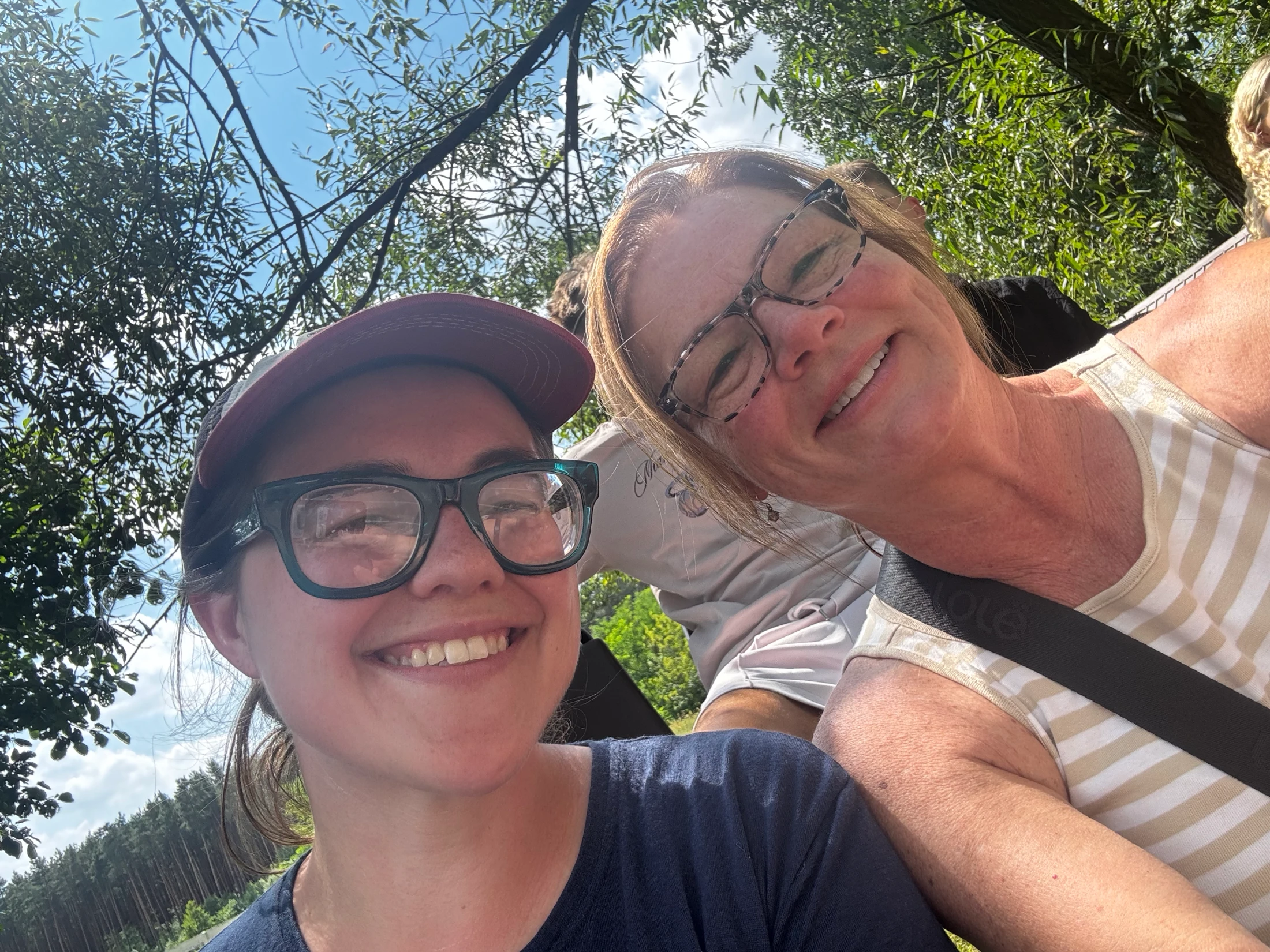 A low-angle selfie of two smiling women outdoors. One wears a red cap and glasses, the other has on a striped tank top. Trees and a blue sky are above them.