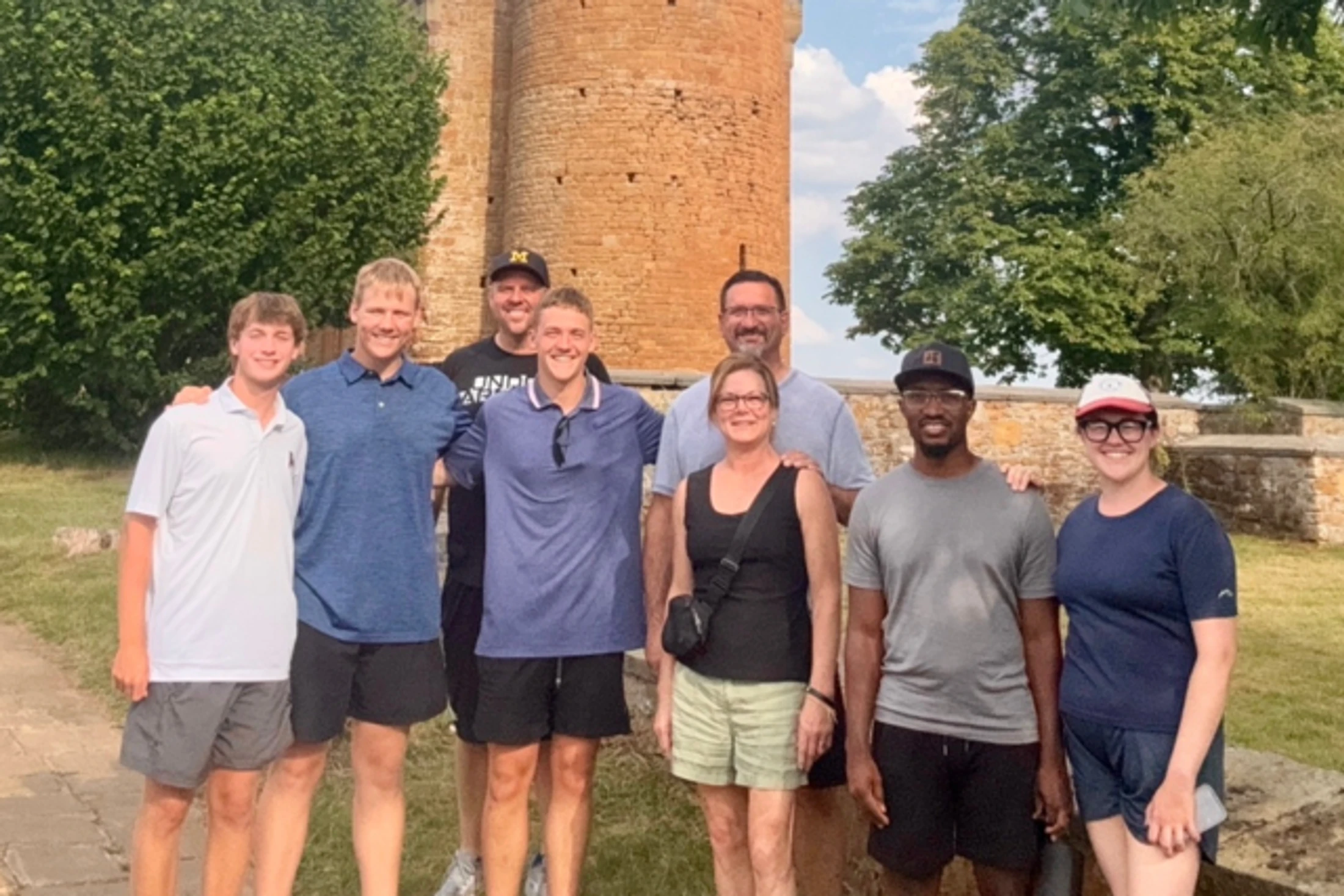 A group of eight smiling people of various ages stand together outdoors for a photo in front of a tall, circular stone tower and lush green trees.
