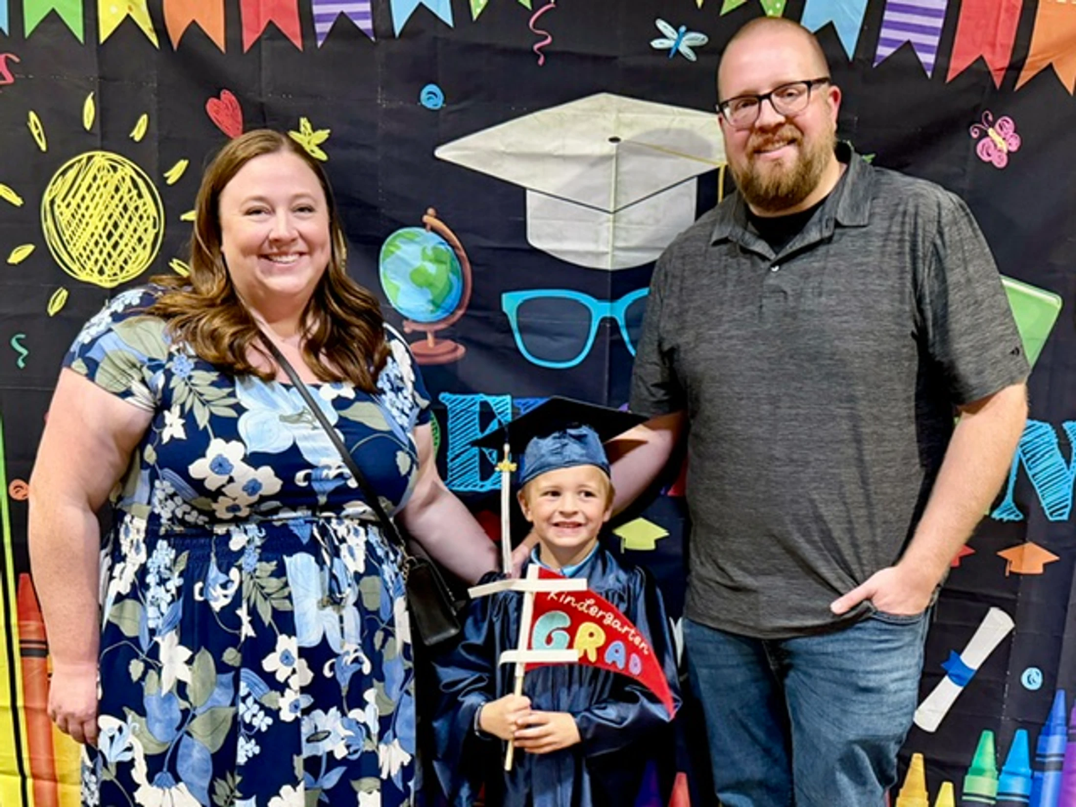 A smiling family of three poses for a photo at a kindergarten graduation. The young boy wears a cap and gown, standing between his proud parents.