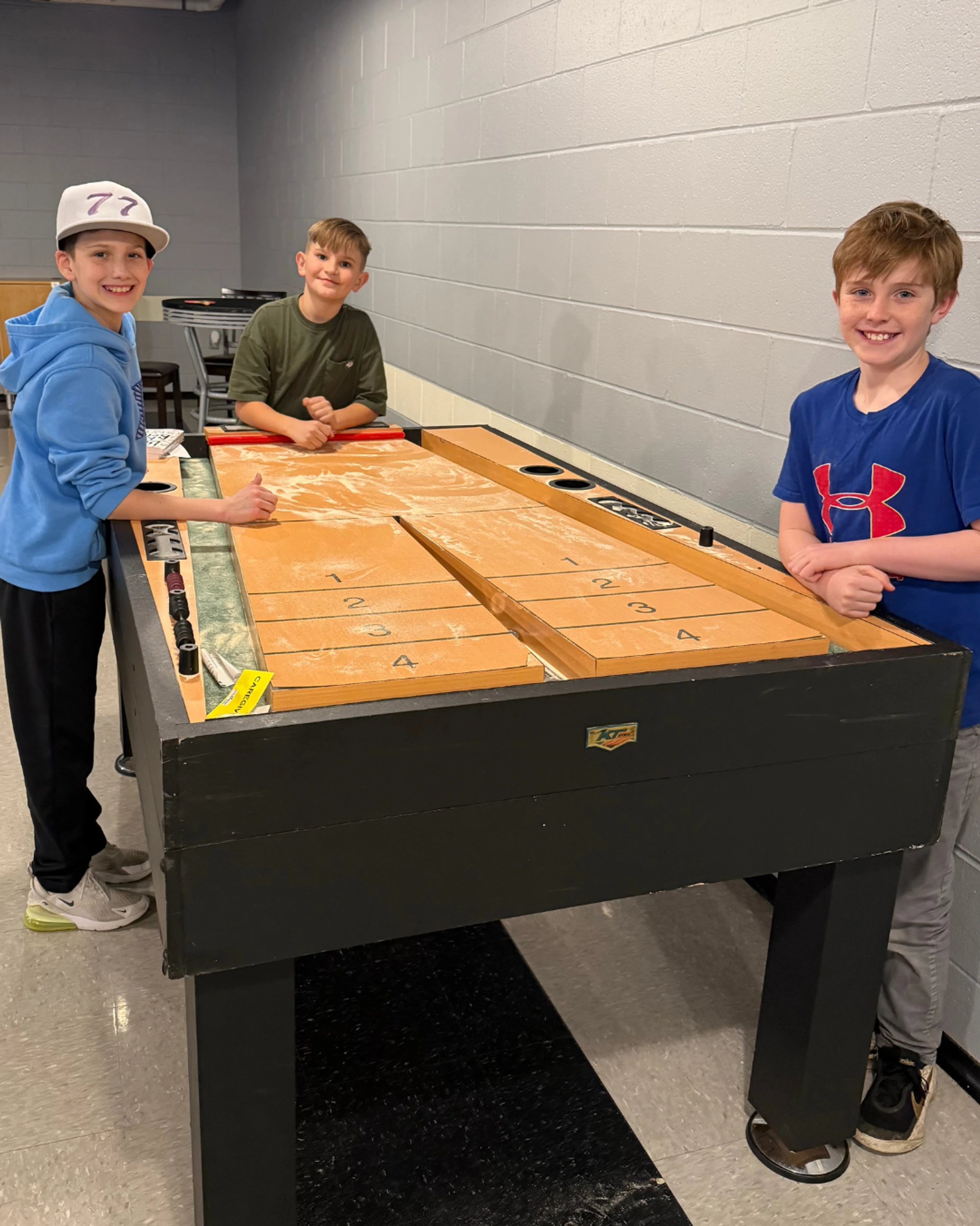 Three young boys smile for the camera while standing around a wooden shuffleboard table in a room with grey block walls.