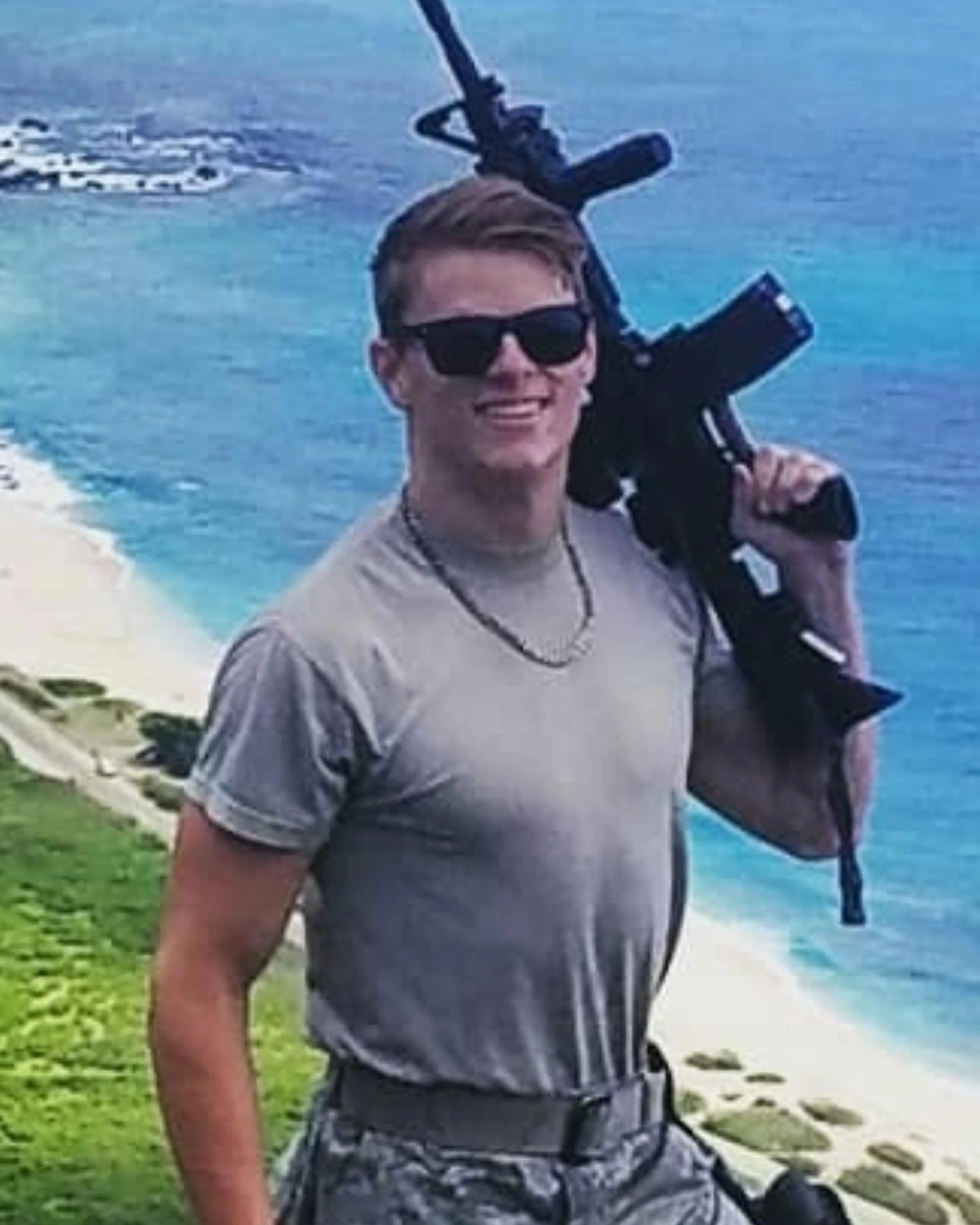 A young man in military attire and sunglasses smiles while carrying a rifle over his shoulder, with a scenic view of a tropical beach and ocean behind him.