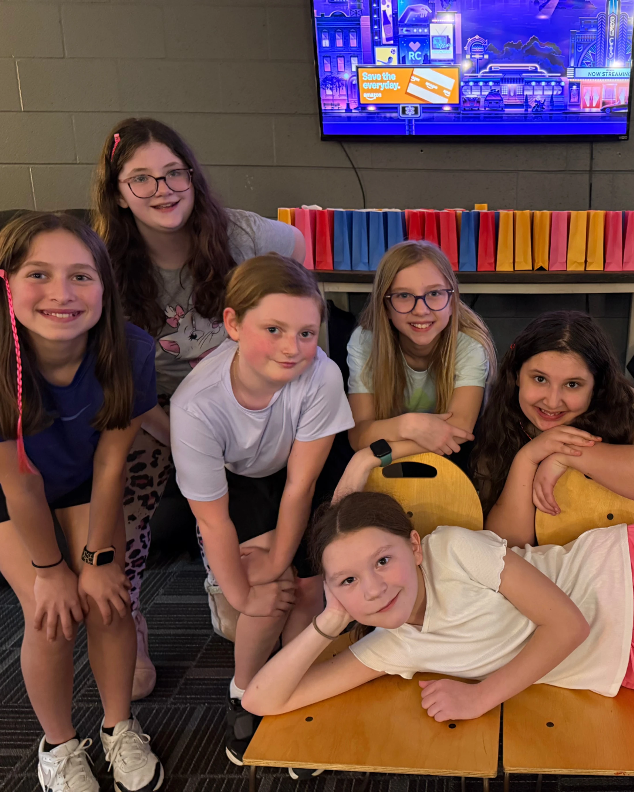Six young girls smiling and posing together for a group photo. In the background are colorful party favor bags and a large television screen.