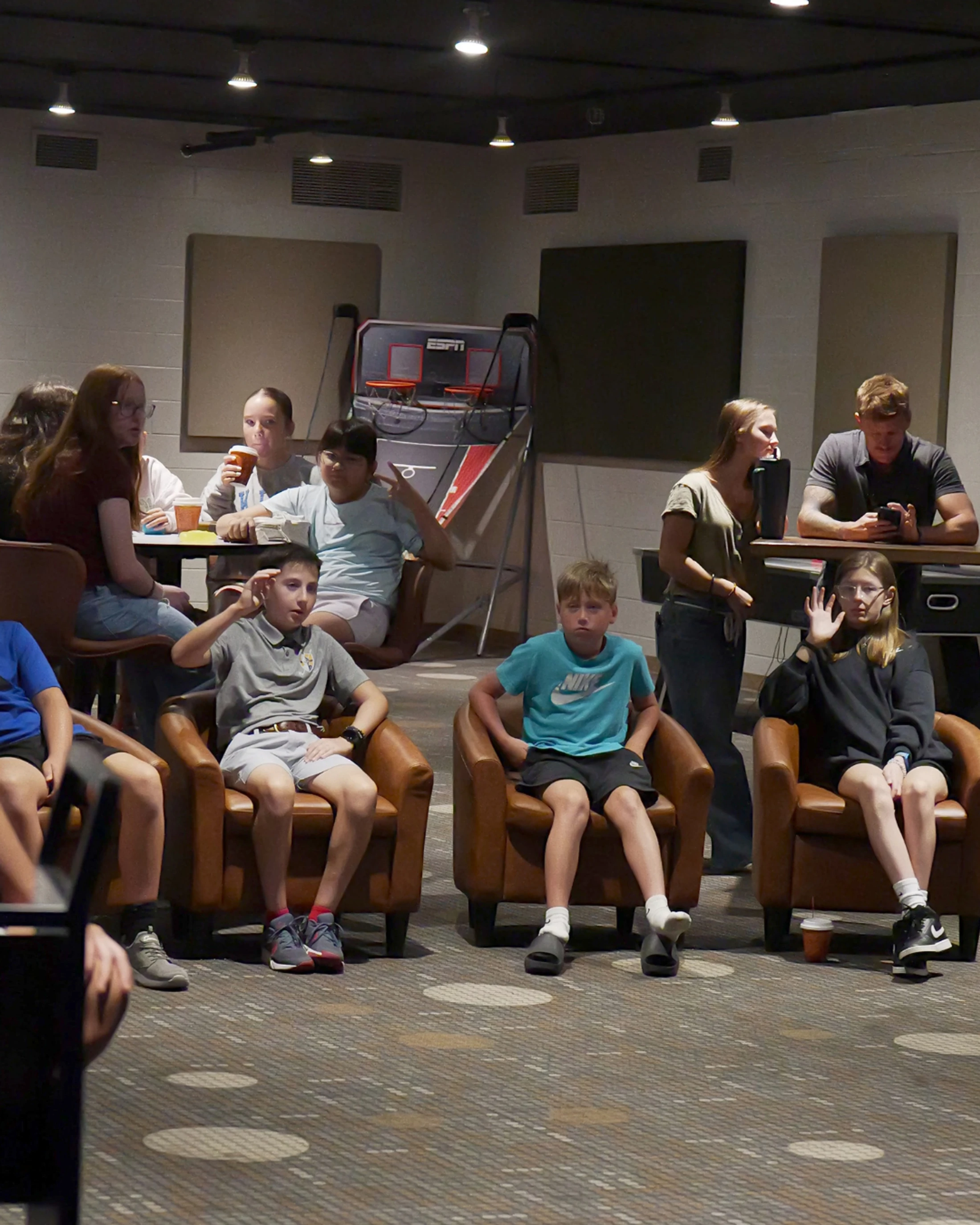 A group of kids and teenagers sit in brown leather chairs and at tables in a modern common room, listening and interacting during a gathering.