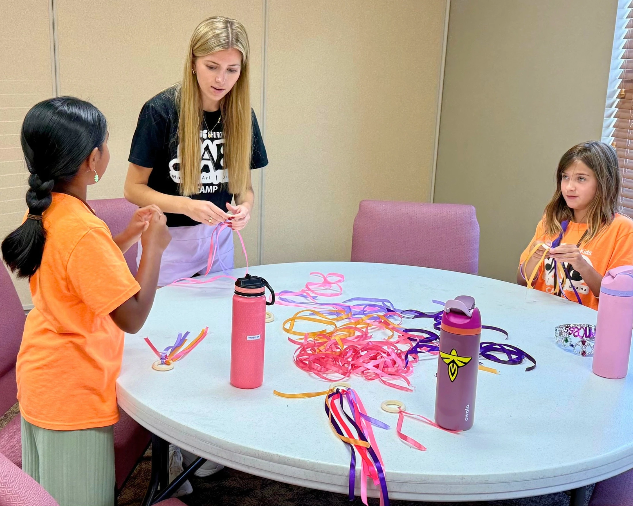 A female camp counselor helps two young girls make crafts by tying colorful ribbons to wooden rings while sitting at a round white table.