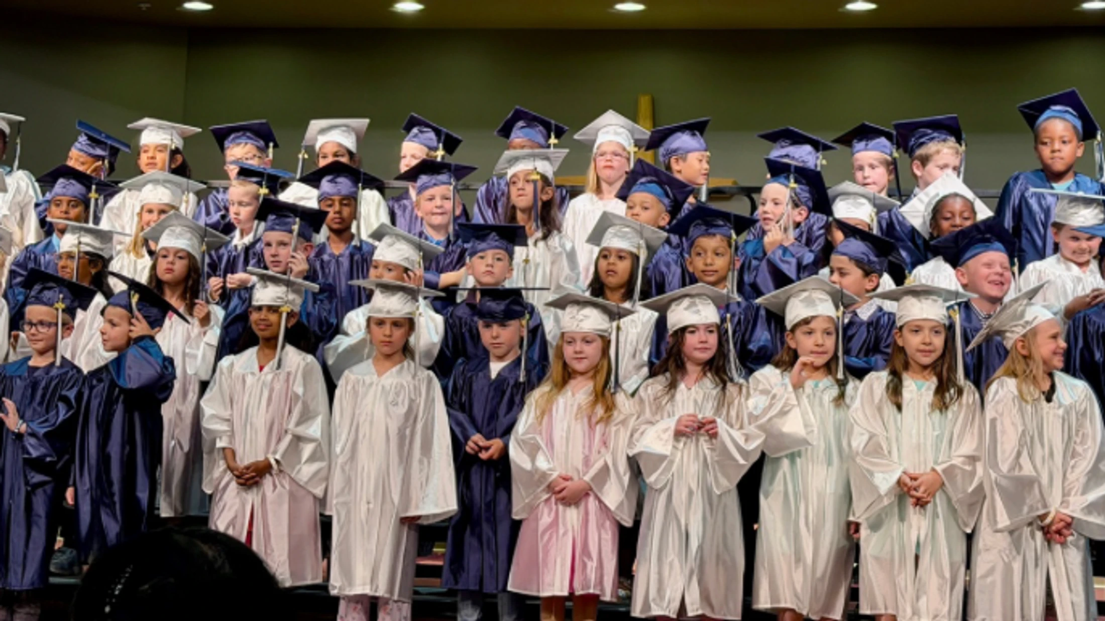 A large group of diverse young children wearing blue and white graduation caps and gowns stand together on risers for a school ceremony.