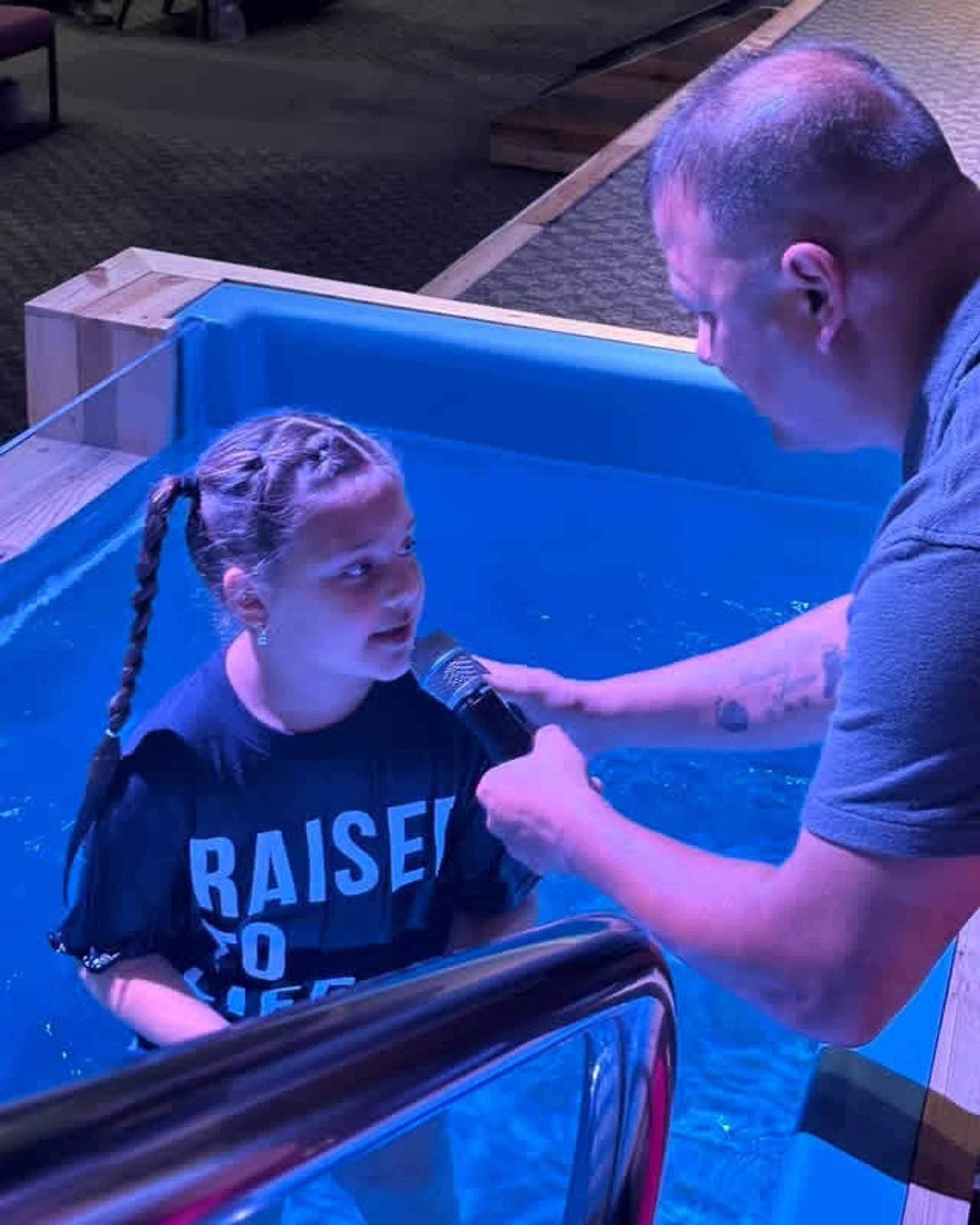 A young girl with braided pigtails stands in a blue baptismal pool, speaking into a microphone held by a pastor before her immersion.