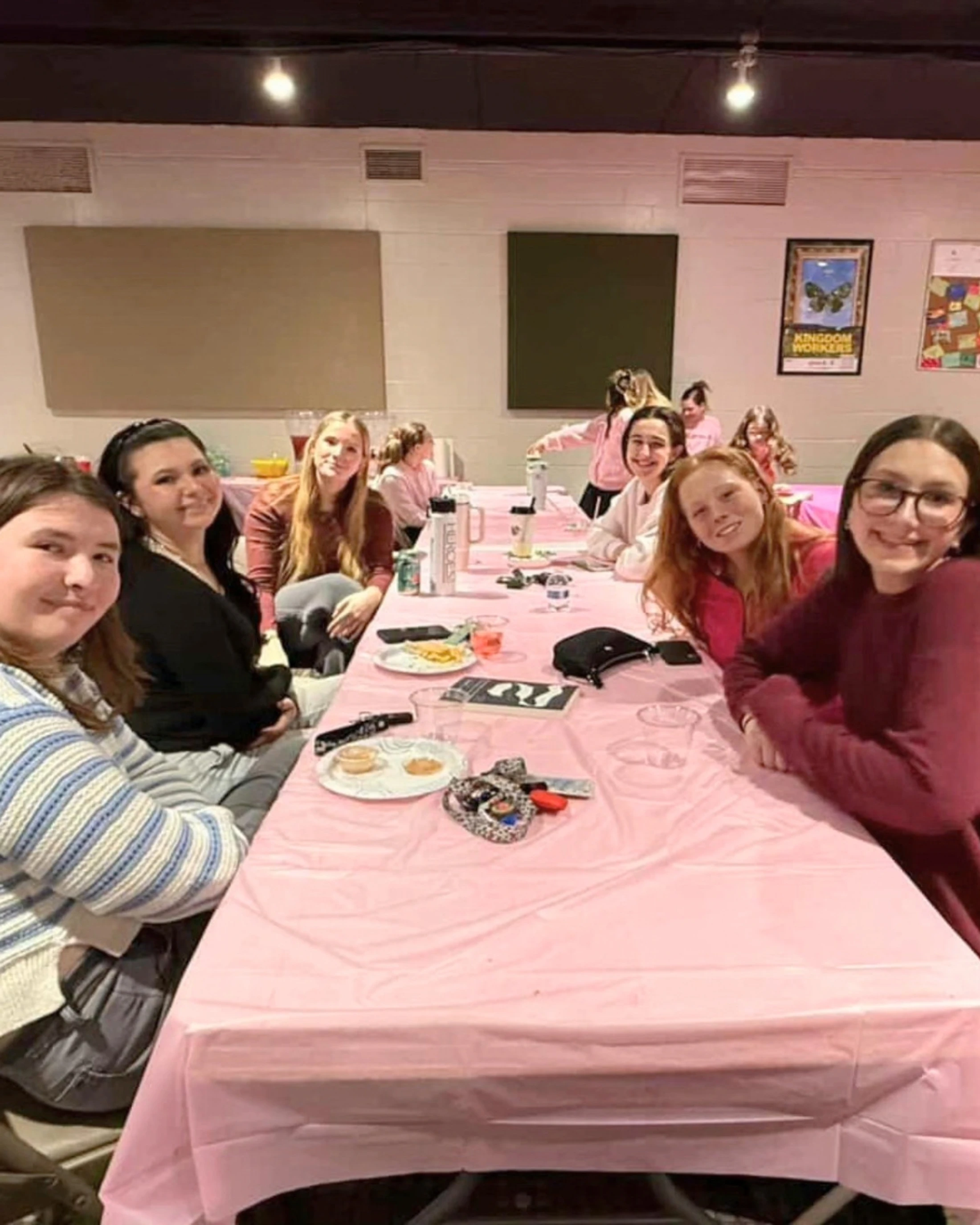 A group of young women and teenage girls smile for the camera while seated at a long table covered with a pink plastic tablecloth during an indoor event.