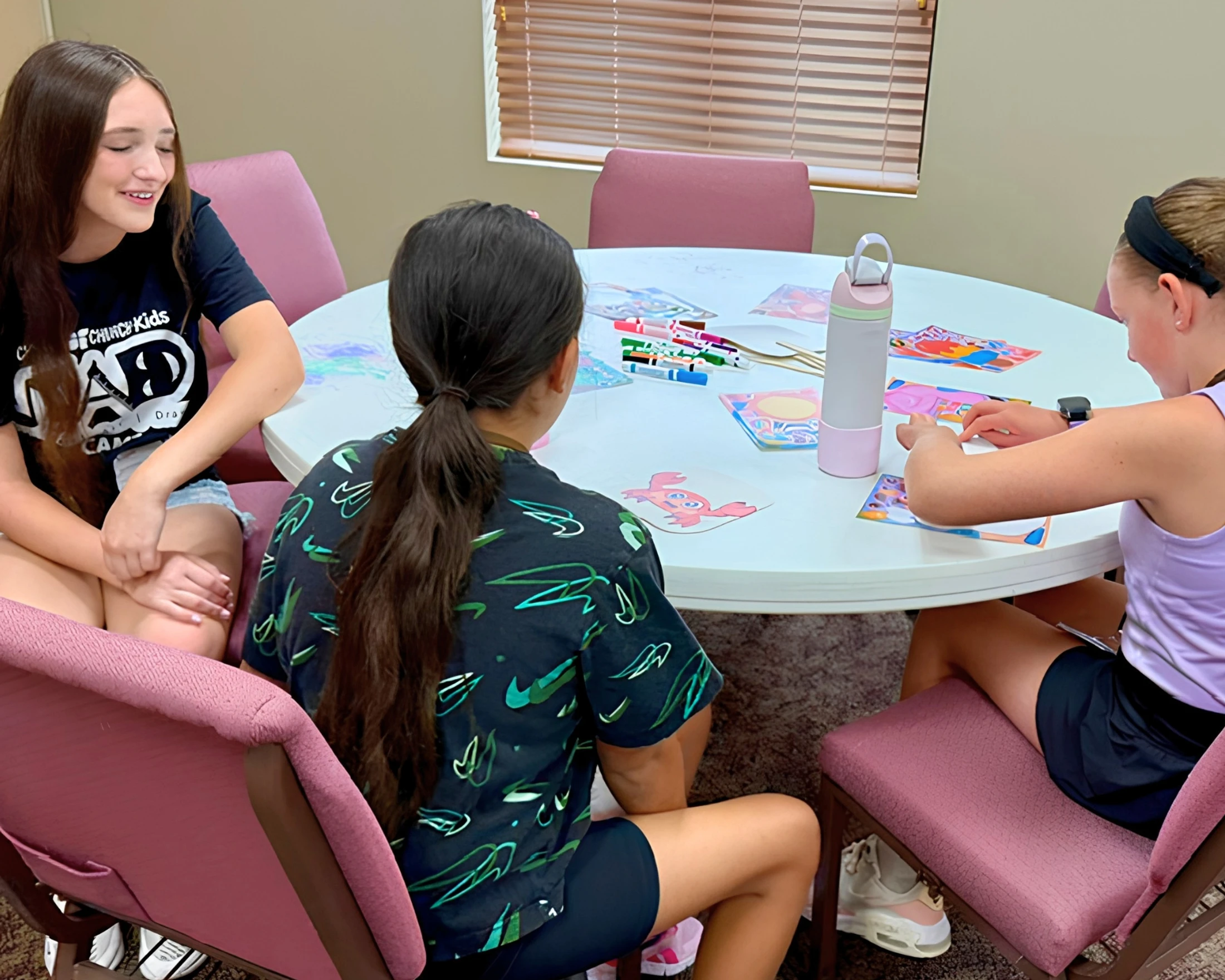 Three young girls sit around a white circular table, engaged in an arts and crafts activity with markers and paper cutouts.