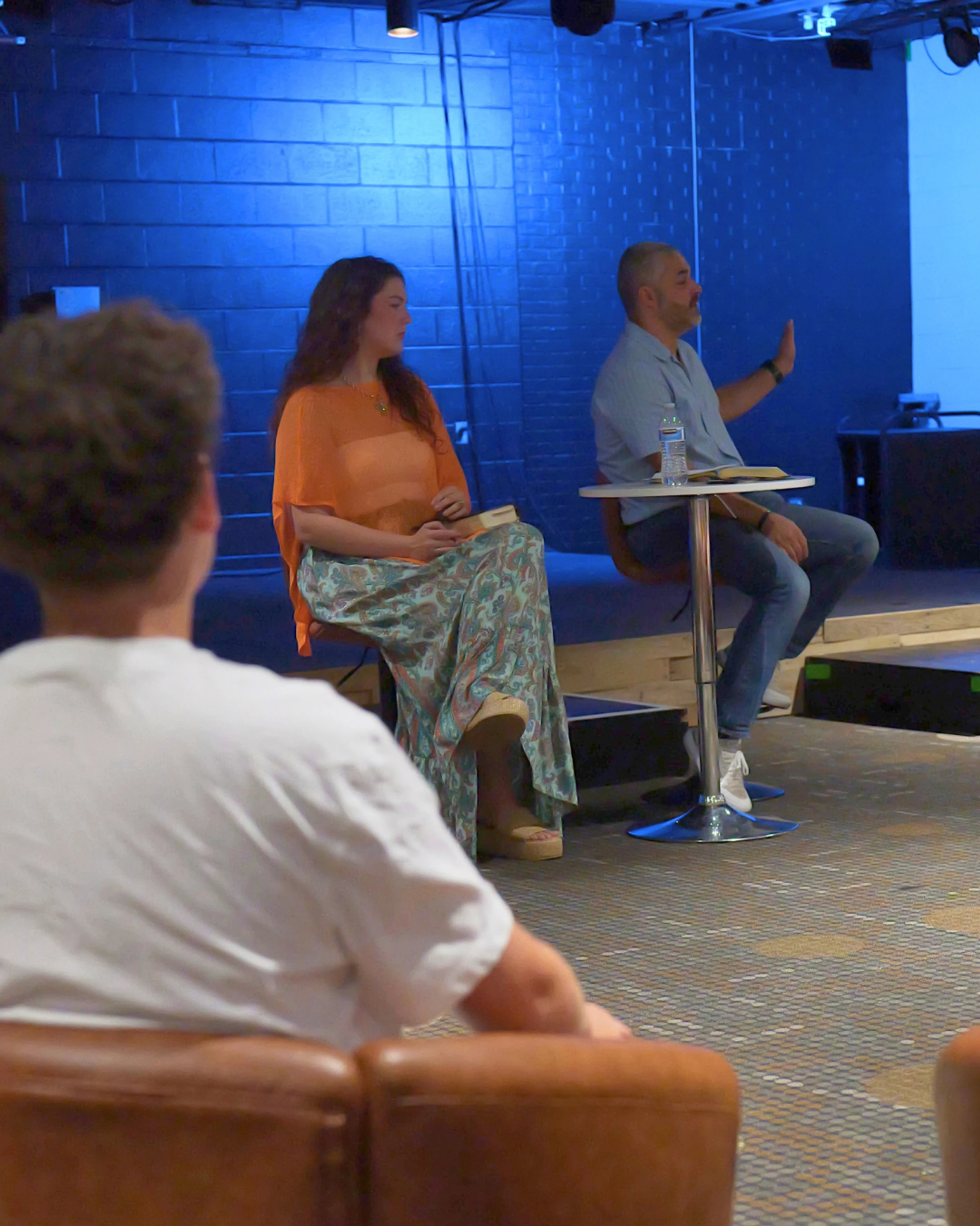 A man and a woman sit on a stage with a blue-lit background, speaking to an audience. The man gestures while talking, and the woman listens attentively.