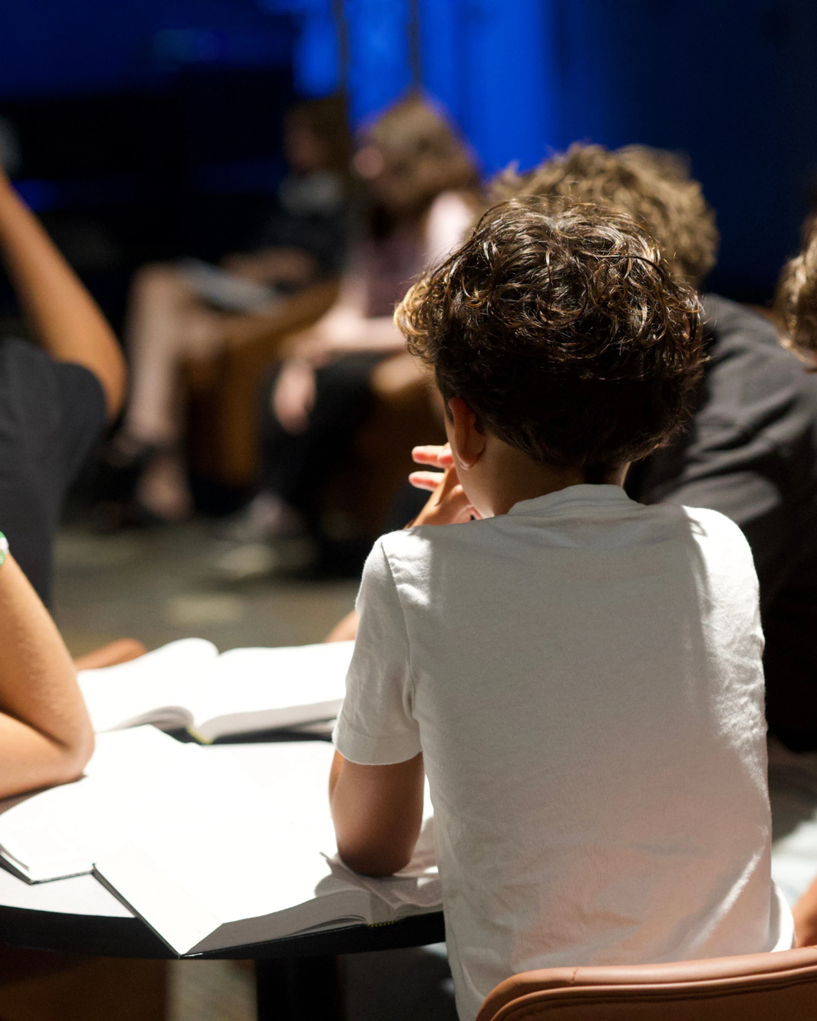 From behind, a young person with curly hair in a white shirt sits at a table with an open book, attentively listening during a class or lecture.
