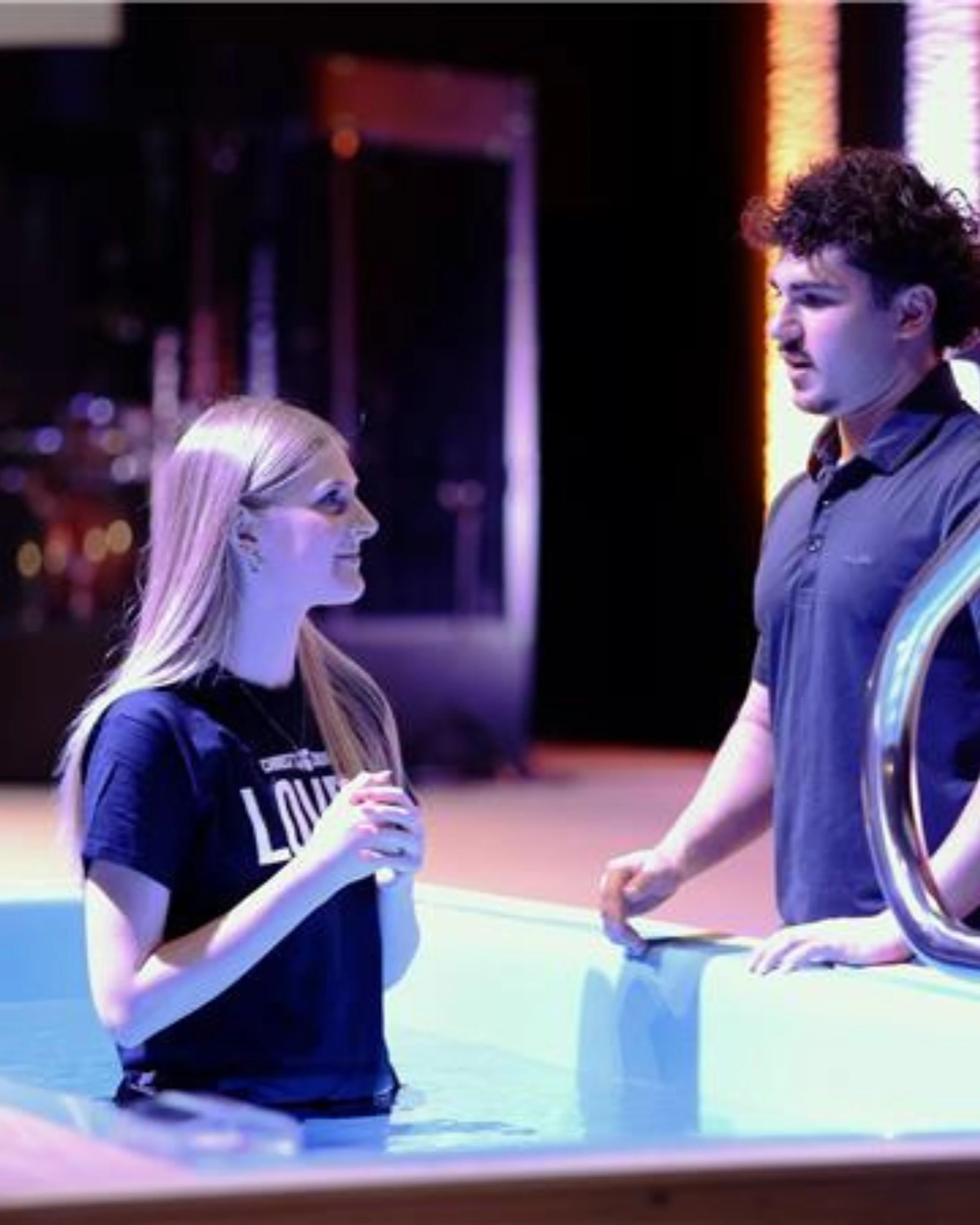 A young woman with blonde hair stands in a baptismal pool, speaking with a young man who is officiating her baptism in a church.