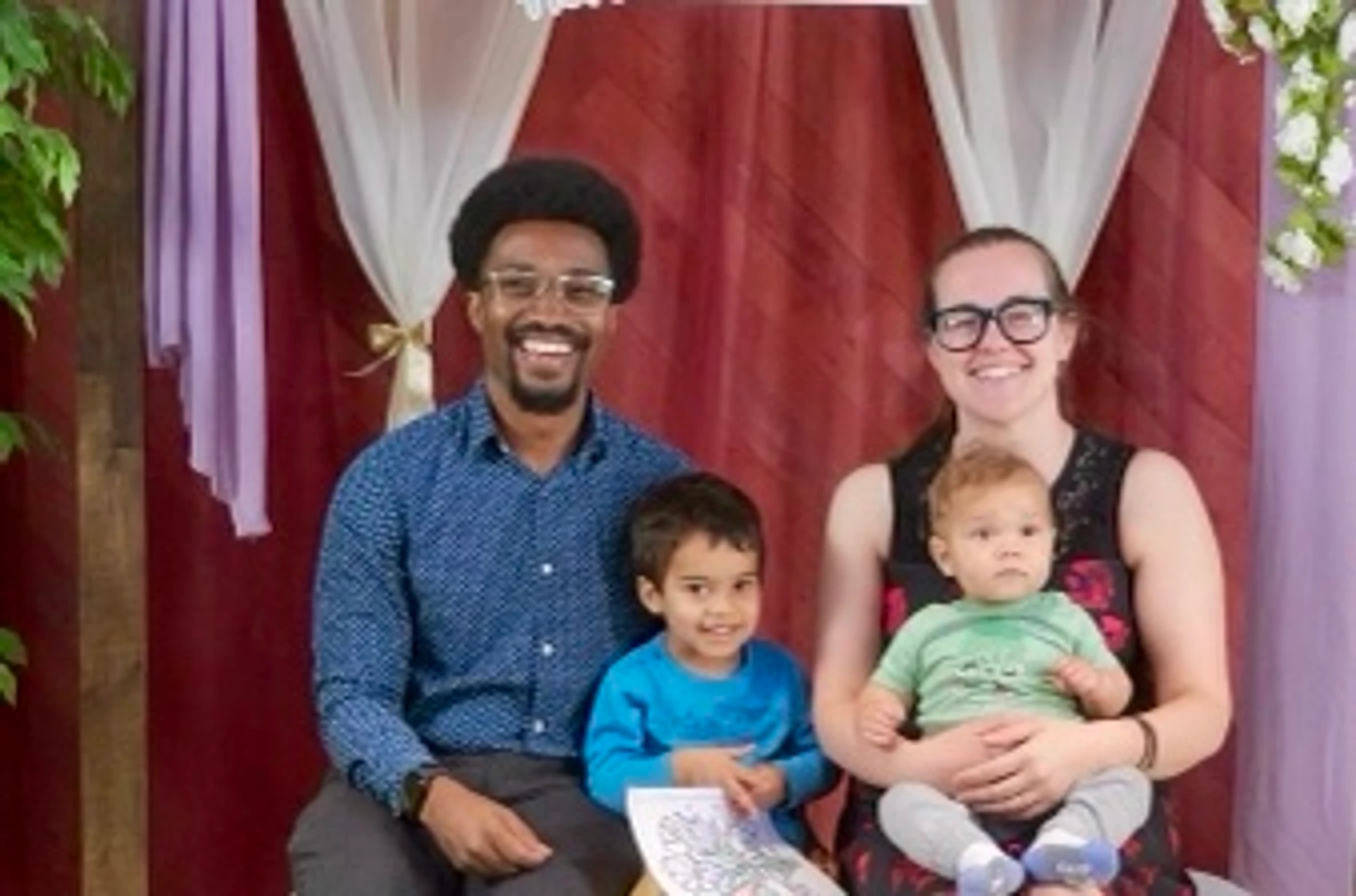 A smiling family of four, a Black man, a white woman, and their two young sons, sit together for a portrait in front of a draped background.