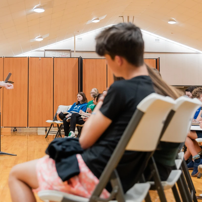 From behind a student, a man in a polo shirt speaks into a microphone to an audience of young people sitting on chairs in a large hall with wood floors.