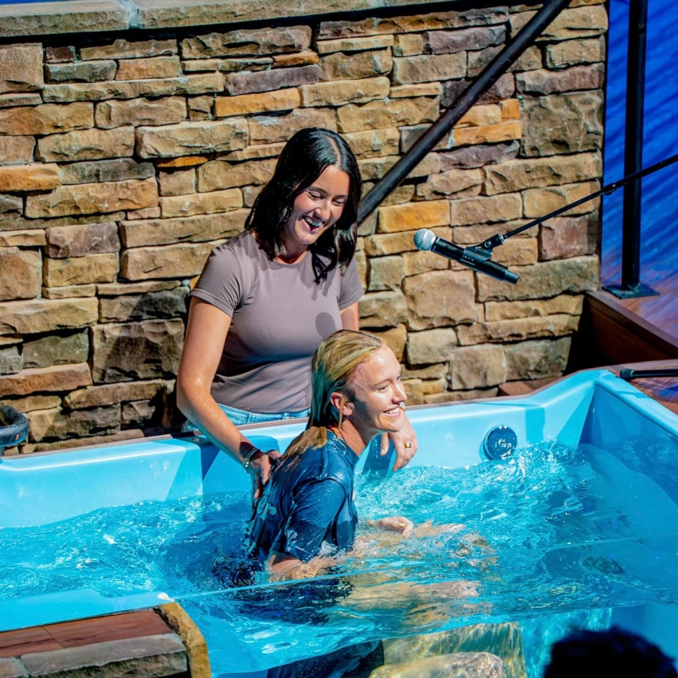 A woman smiles as she prepares to baptize another smiling woman who is sitting in a small blue pool of water in front of a stone wall.