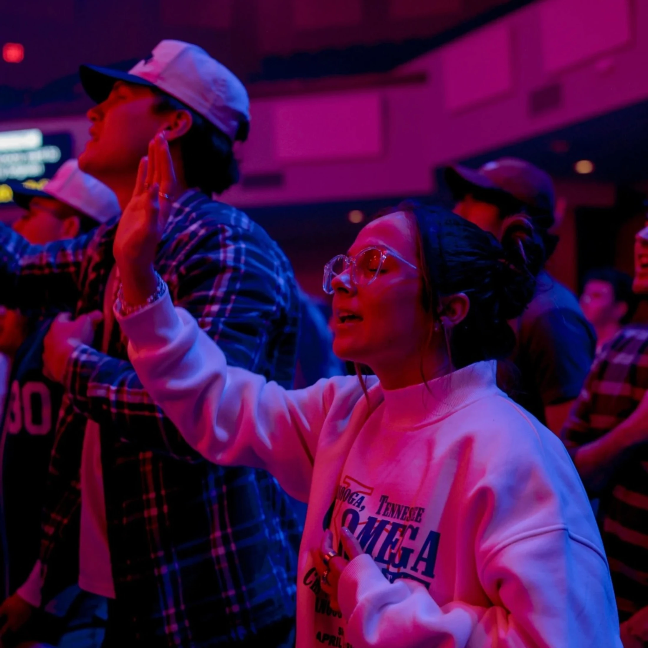 A young woman with glasses raises her hand while singing passionately in a crowd. The scene is illuminated by vibrant pink and blue lights during an event.