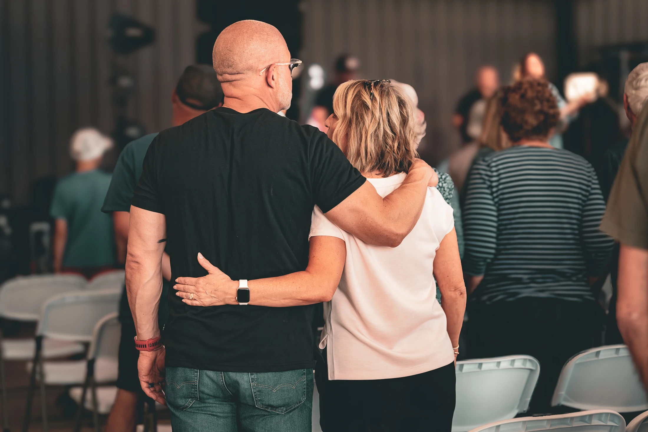 From behind, a bald man in a black shirt has his arm around a woman in a white shirt. They stand together in a crowd of people at an event.