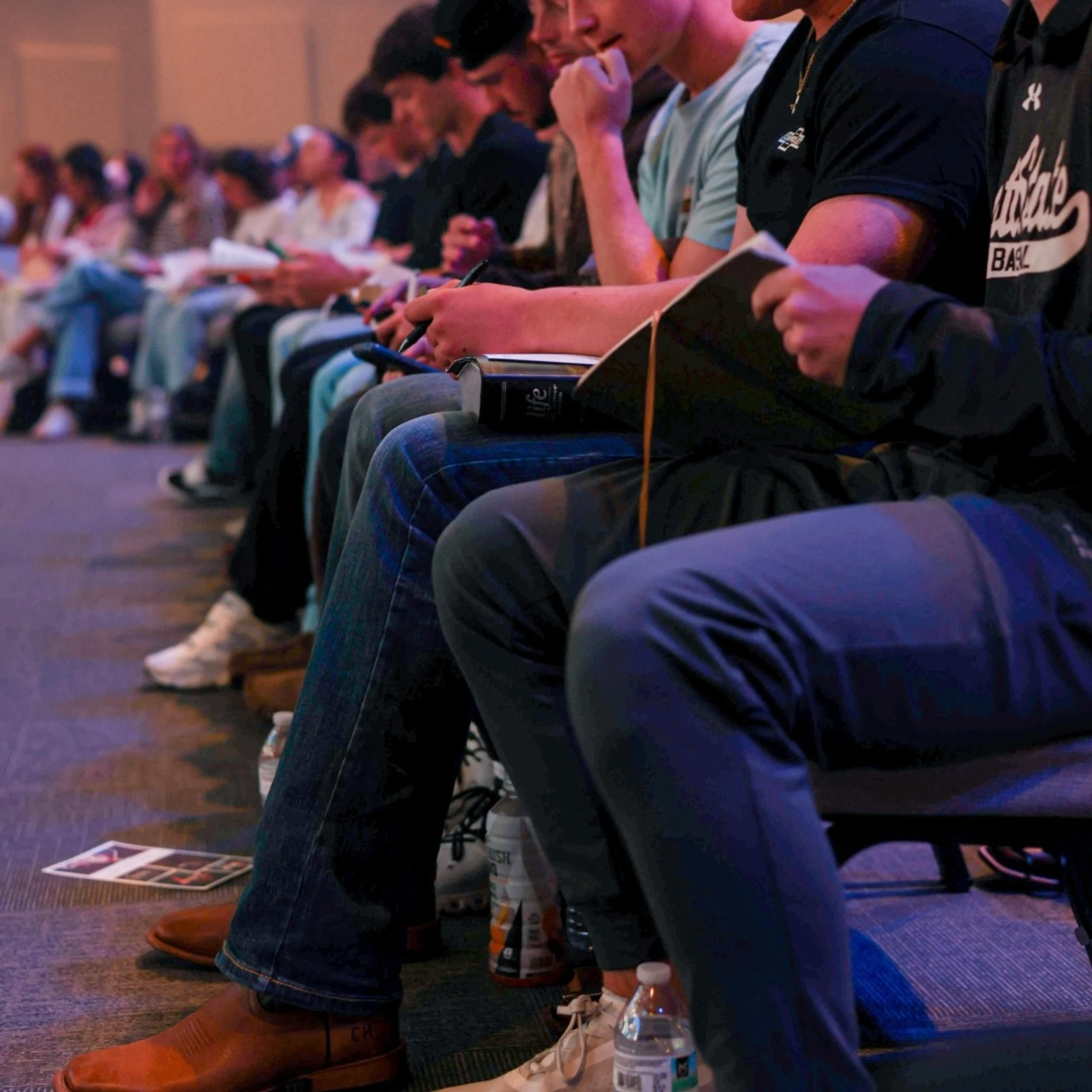 A low-angle view of a row of young adults sitting in chairs, holding notebooks and Bibles on their laps as they listen attentively at a seminar.