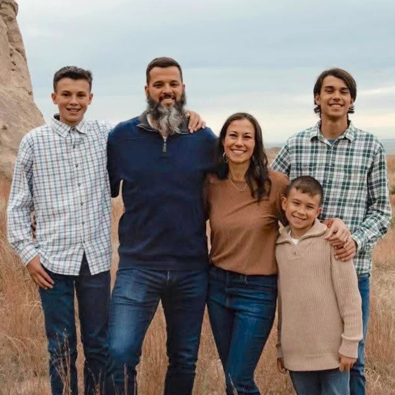 A family of five, two parents and three sons, stand together smiling for a photo in a field of tall grass with a rock formation in the background.