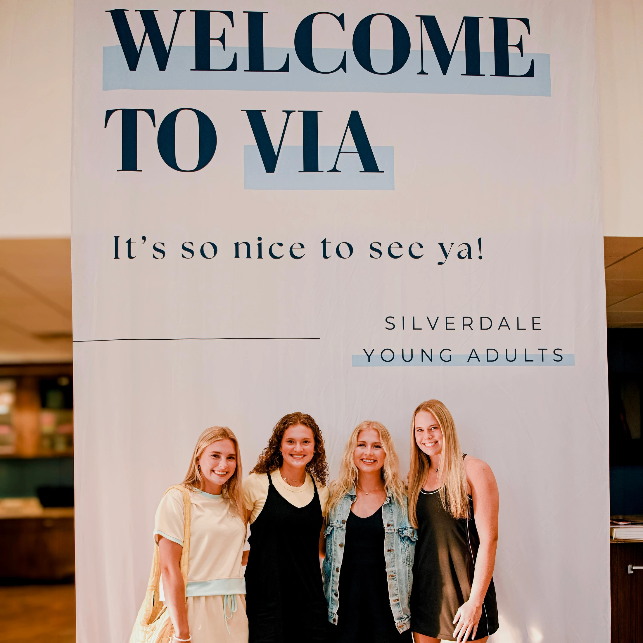 Four young women smile while standing in front of a large banner that reads 'Welcome to VIA' for the Silverdale Young Adults group.