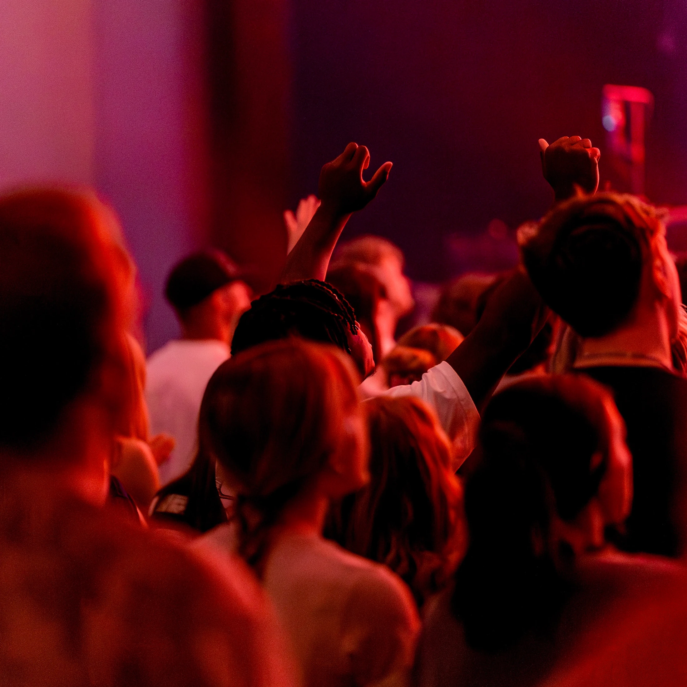 A crowd of people viewed from the back at a concert or event, bathed in atmospheric red light. Some people have their hands raised in worship or celebration.