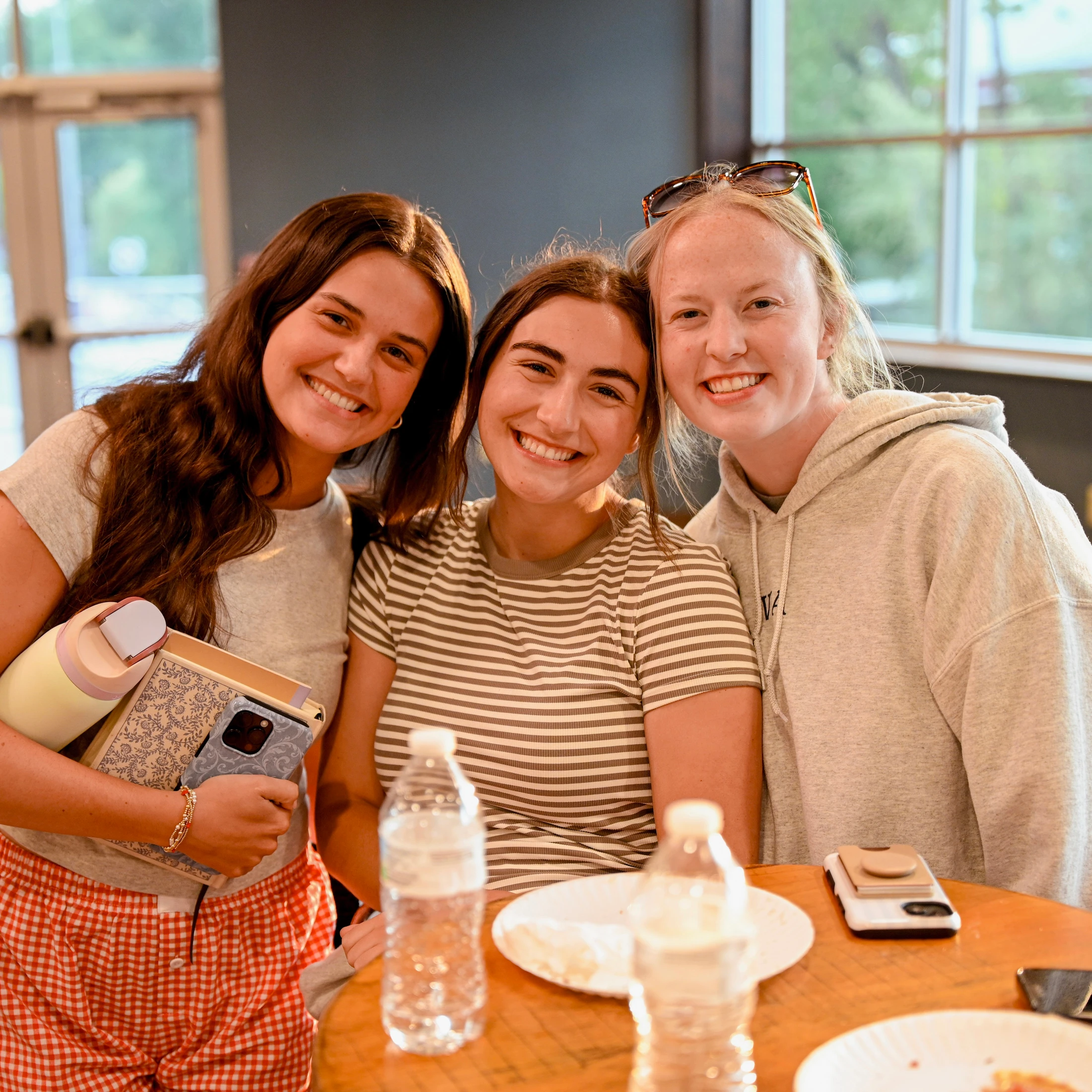 Three young women in casual clothes smile for the camera while gathered around a wooden table with water bottles and paper plates.