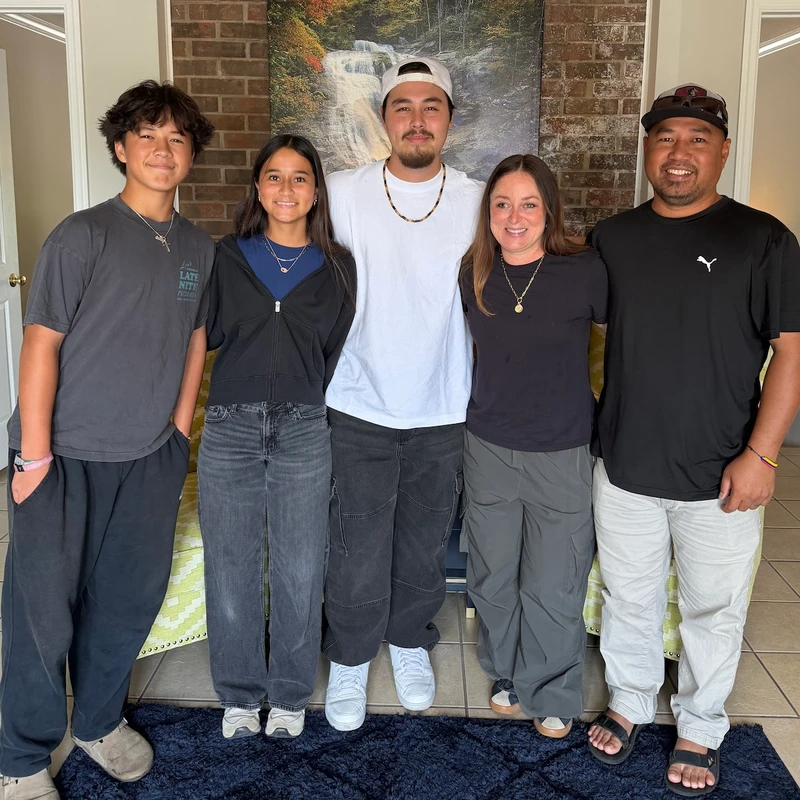 A family of five, two parents and three children, stand side-by-side and smile for a photo in their home in front of a brick wall.