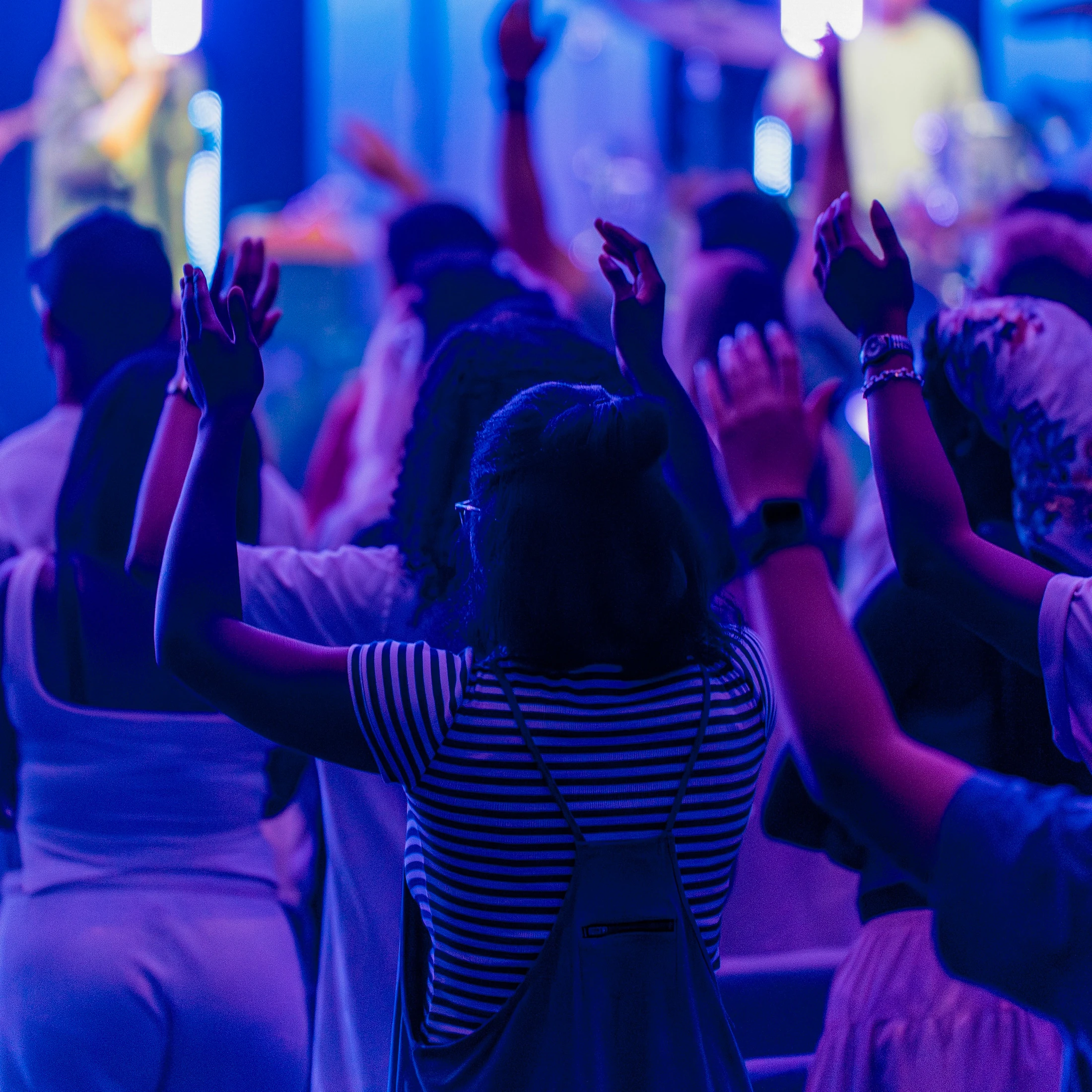 A crowd of people seen from behind with their hands raised in worship. The scene is illuminated with vibrant blue and purple stage lights.