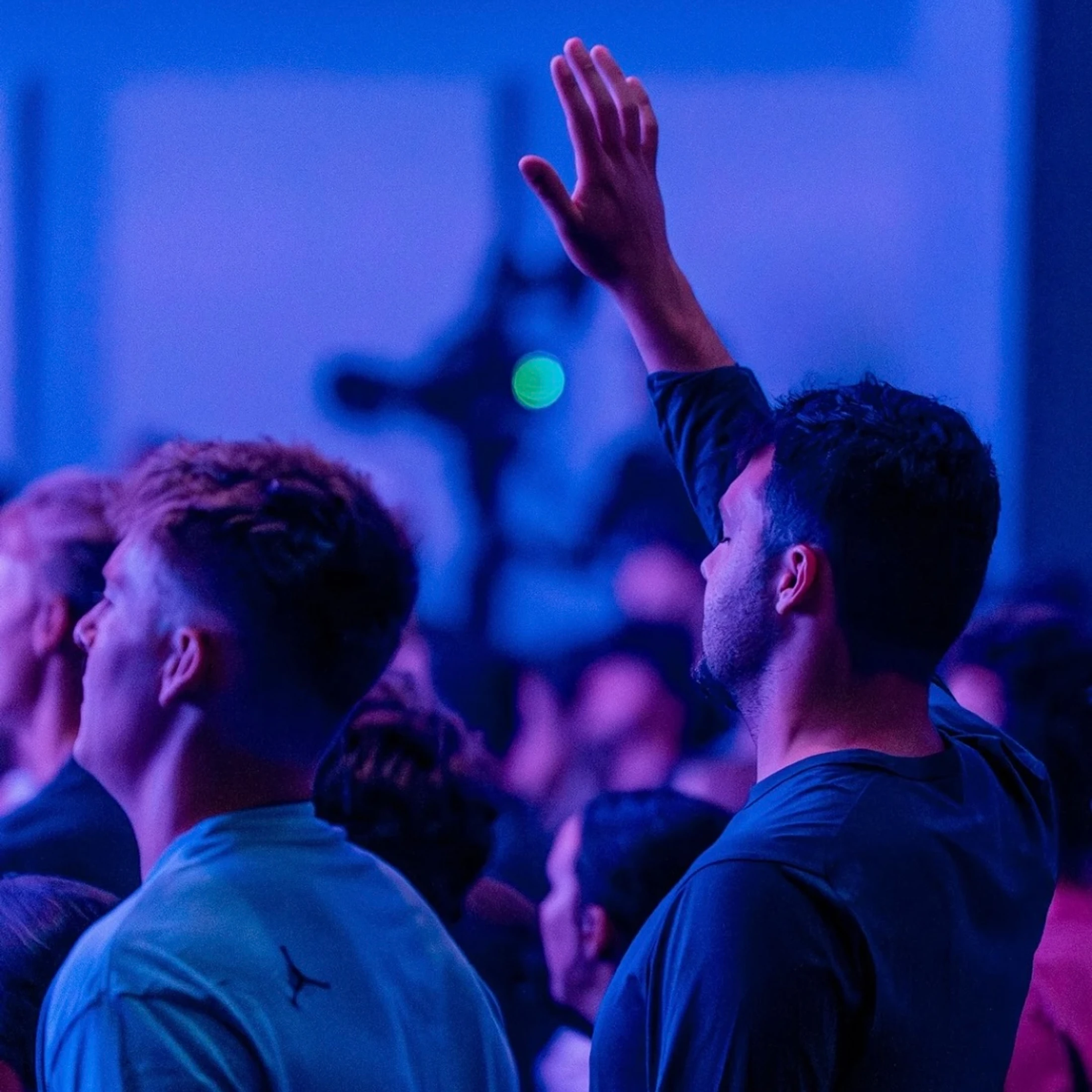 A man raises his hand in a crowd of people bathed in blue and purple light. Another man stands beside him, both looking up towards a stage.